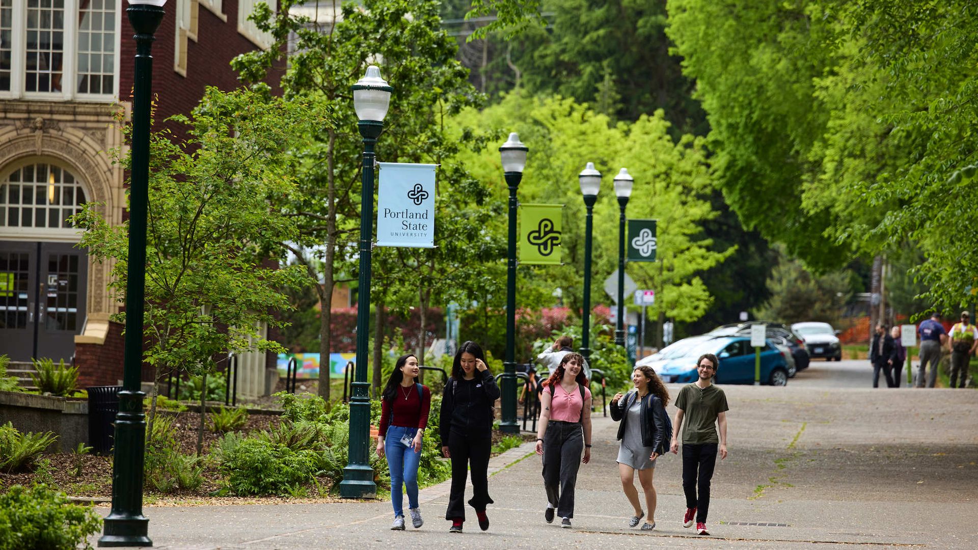 Five diverse students walk and chat on a tree-lined path at Portland State University campus with campus banners and a brick building nearby under overcast skies.