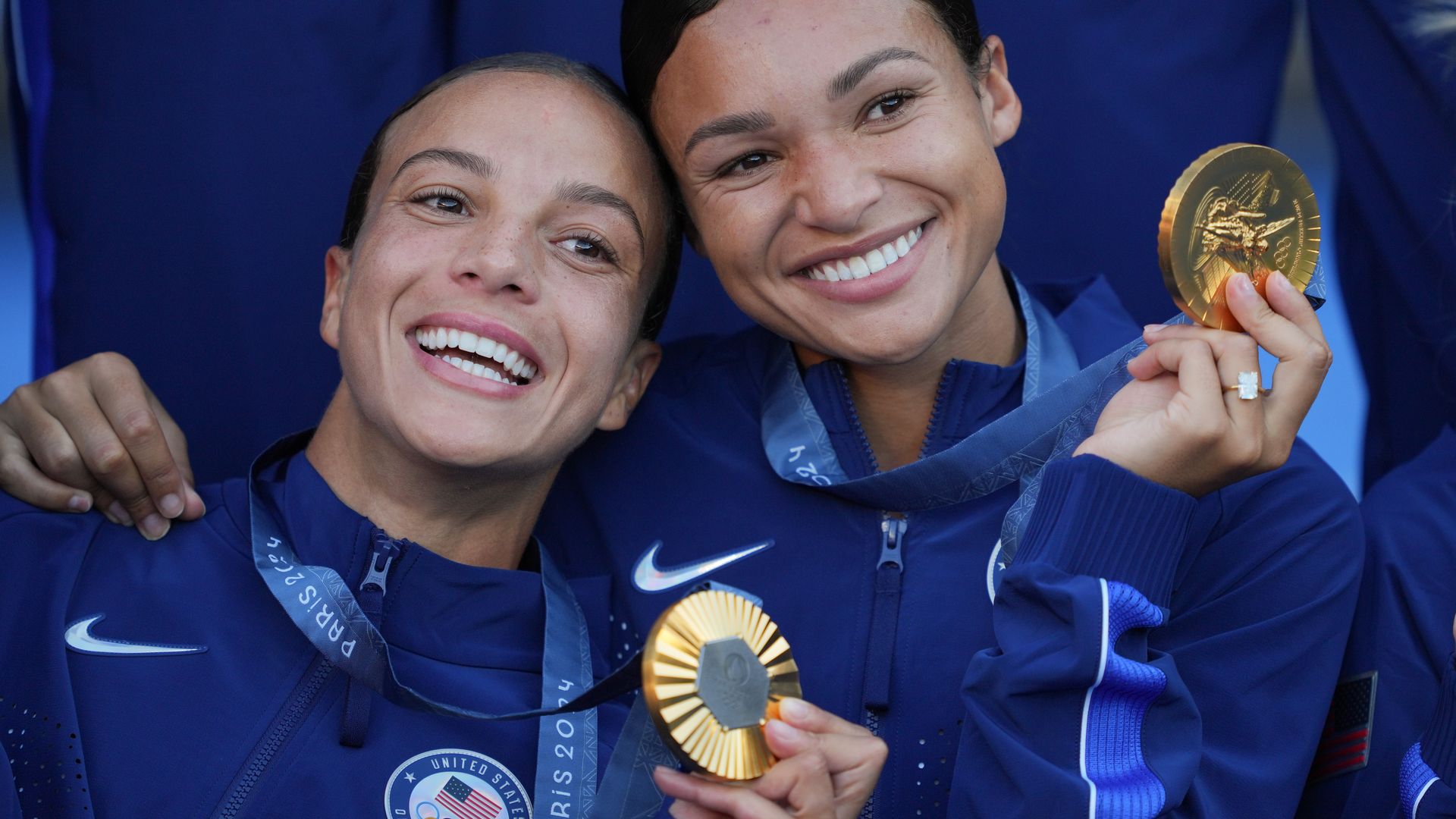 Two women hold up gold medals while smiling. 