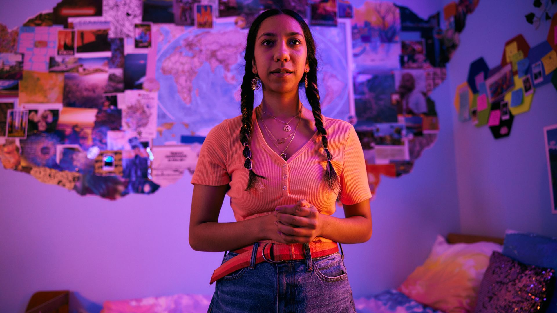 A young woman with two braids stands in a purple-lit room, wearing a pink ribbed top and jeans with a red plaid belt. The wall behind her is covered with maps, photos, and notes.