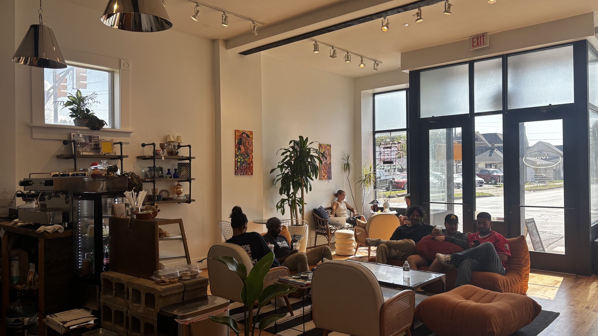 Cozy coffee shop interior with warm lighting, orange and beige chairs, plants, wall art, and several people chatting and relaxing near large front windows showing a sunny street outside.