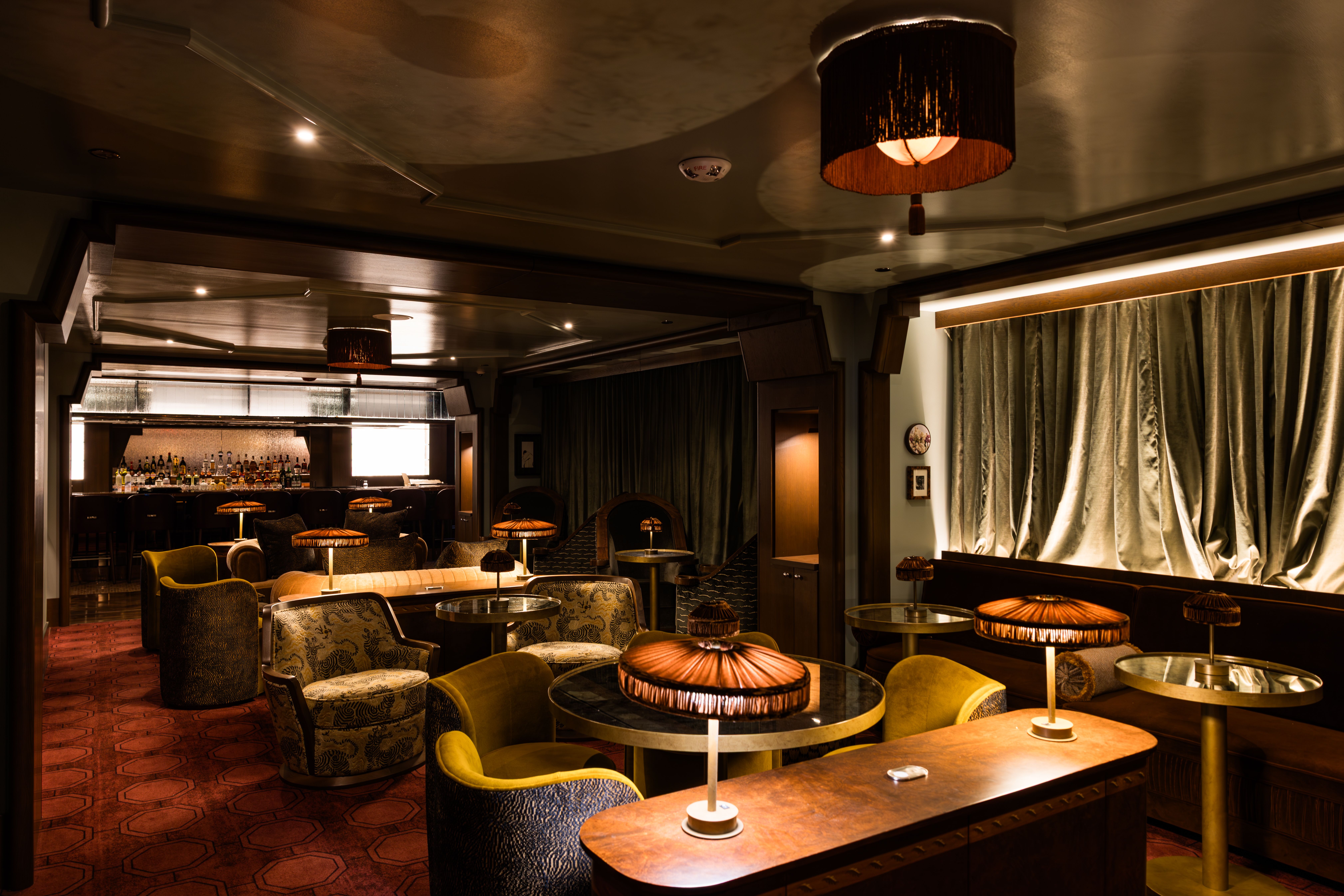 Dim, cozy lounge bar bathed in warm amber light. Round tables with patterned chairs, plush green seating, and heavy curtains. A dark wooden back bar with bottles adds a vintage vibe.