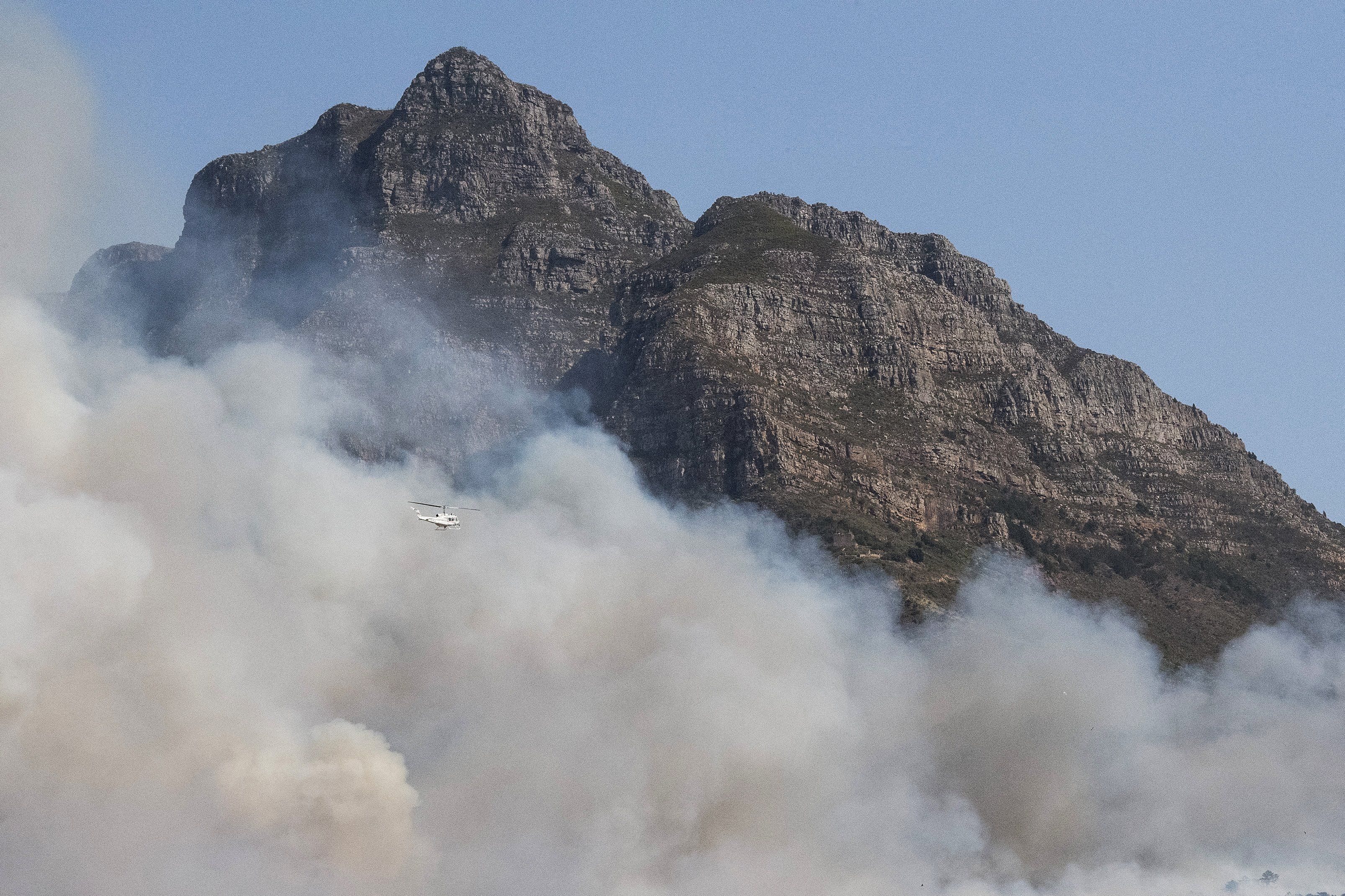 A helicopter flies near a smoke cloud due to a forest fire burning out of control on the foothills of Table Mountain in Cape Town, on April 18