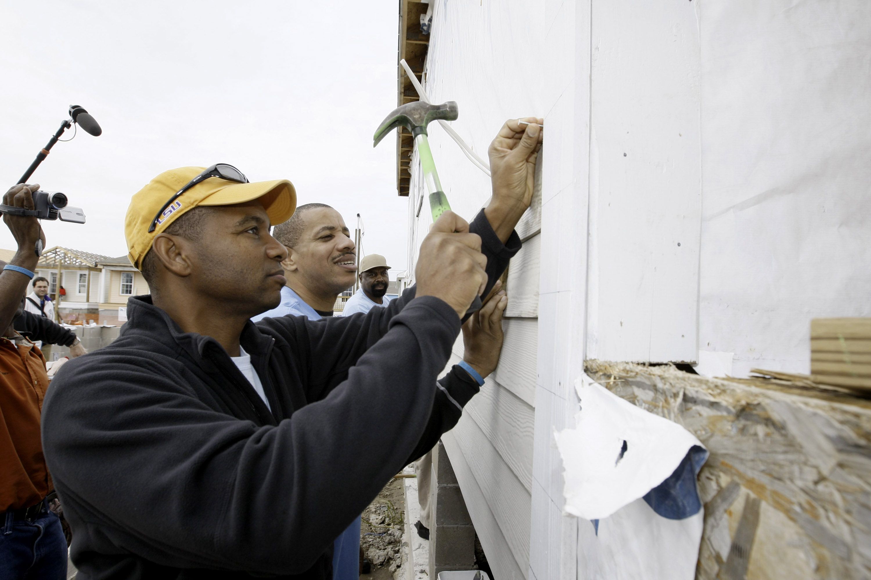 A person positions a nail on the side of a house while others do home improvement on the same property.