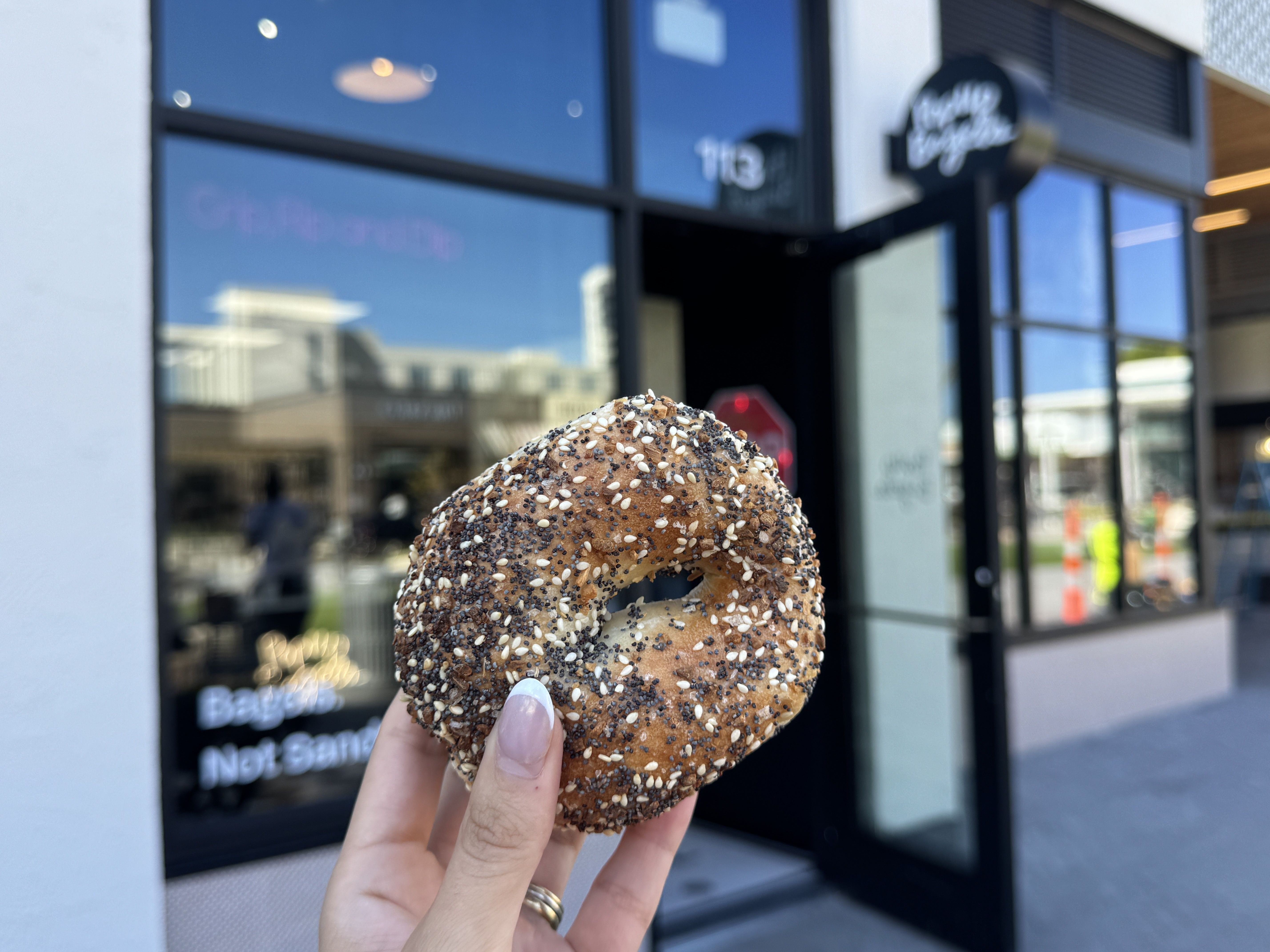 Hand holding a seeded bagel with sesame, poppy, and other seeds in front of a cafe entrance with glass doors and windows under blue sky.