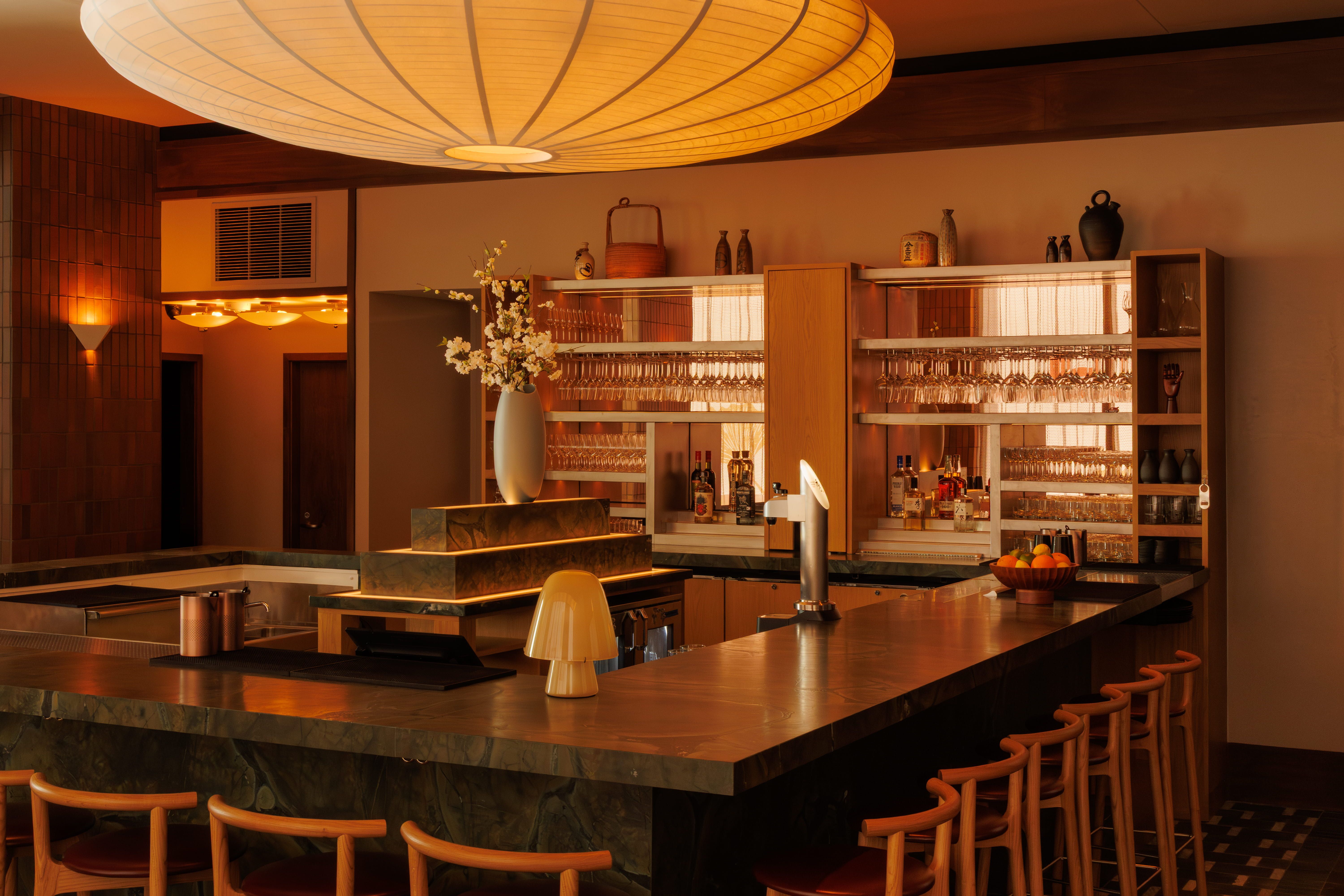 Warm, orange-lit bar interior with a large round lantern overhead. Glassware and bottles line the back shelves; a vase of white blossoms on the counter, a bowl of oranges nearby, and marble countertops.