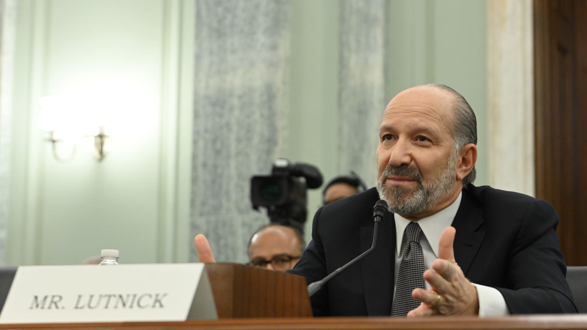 A person who is seated and wearing a suit and tie gestures in front of a microphone