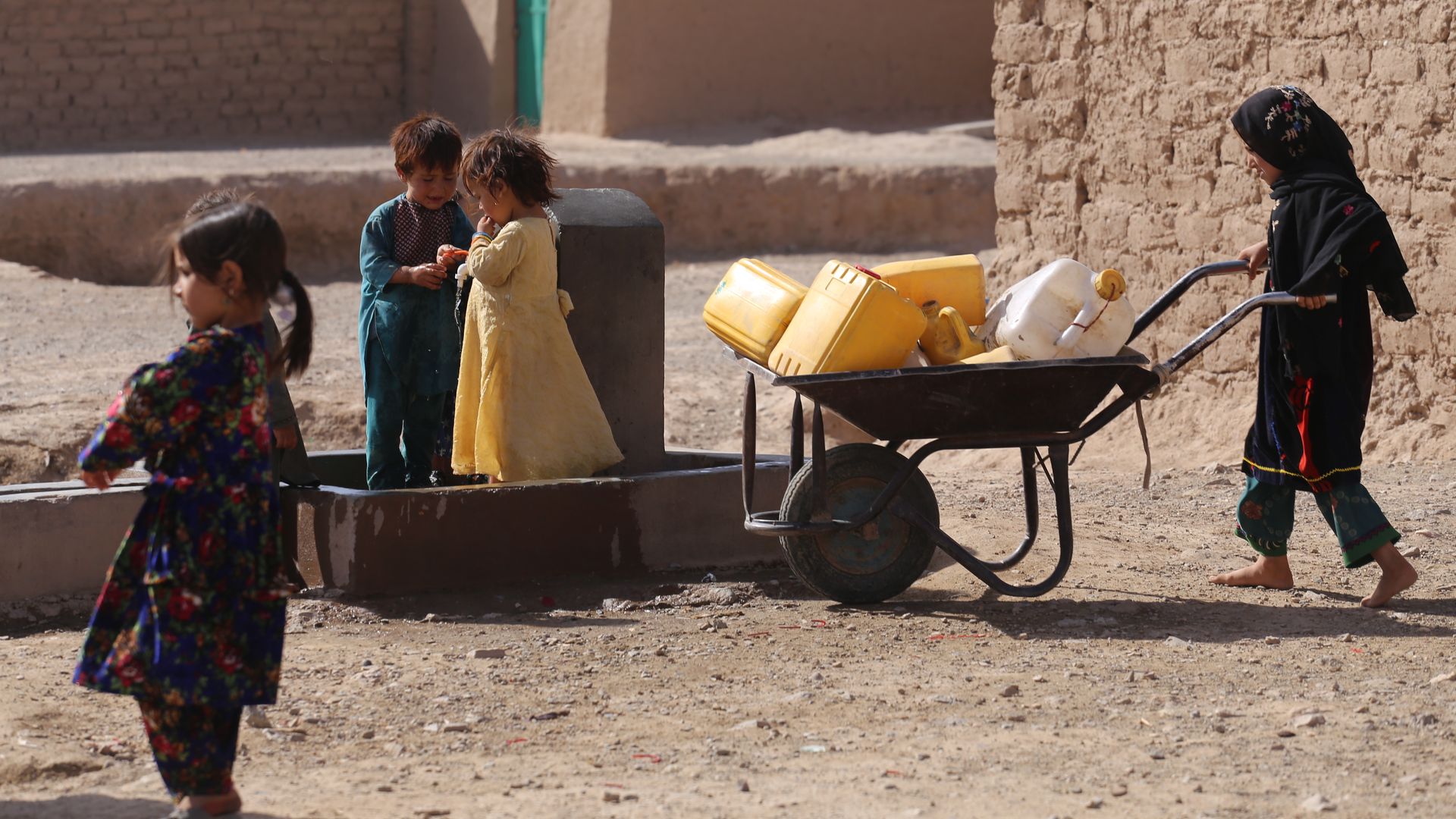 small children in a refugee camp, one pushing a wheelbarrow with empty plastic jugs