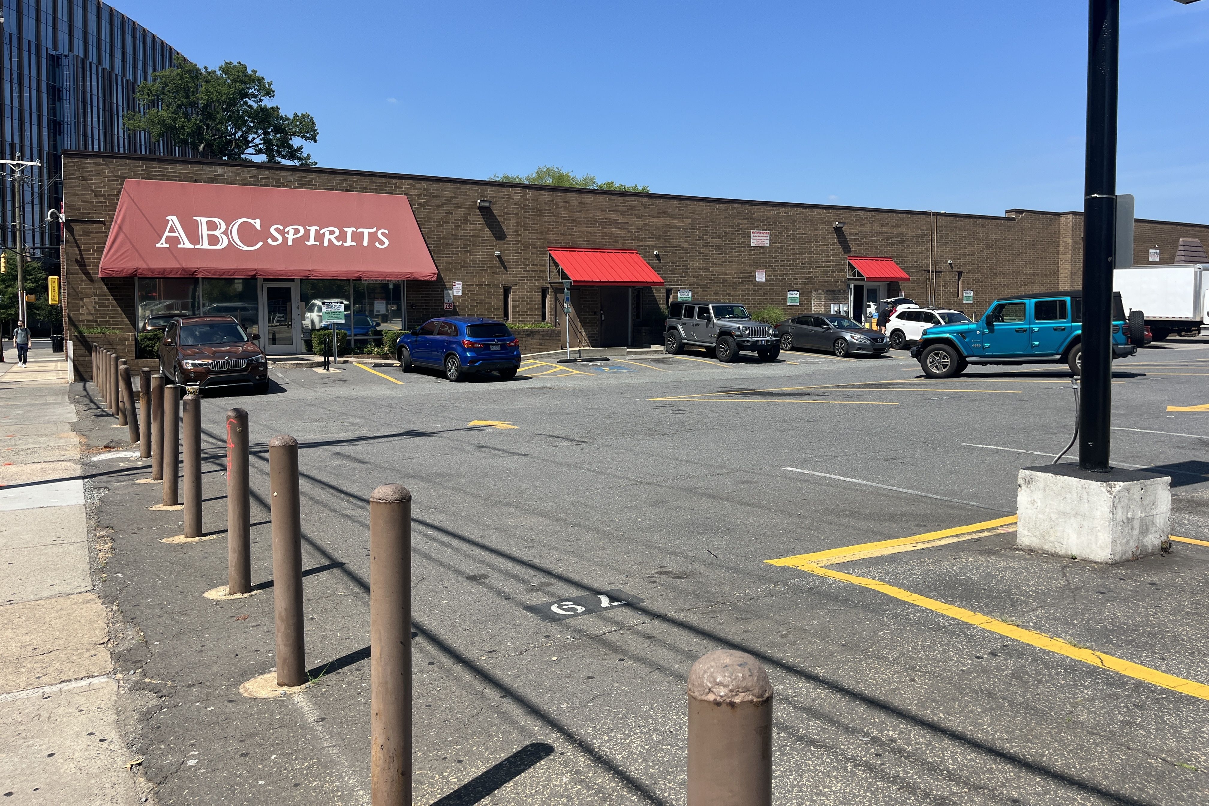 Brick storefront with a maroon awning that reads "ABC SPIRITS" in a parking lot. Several cars, red bay awnings, brown bollards, and a bright blue sky.