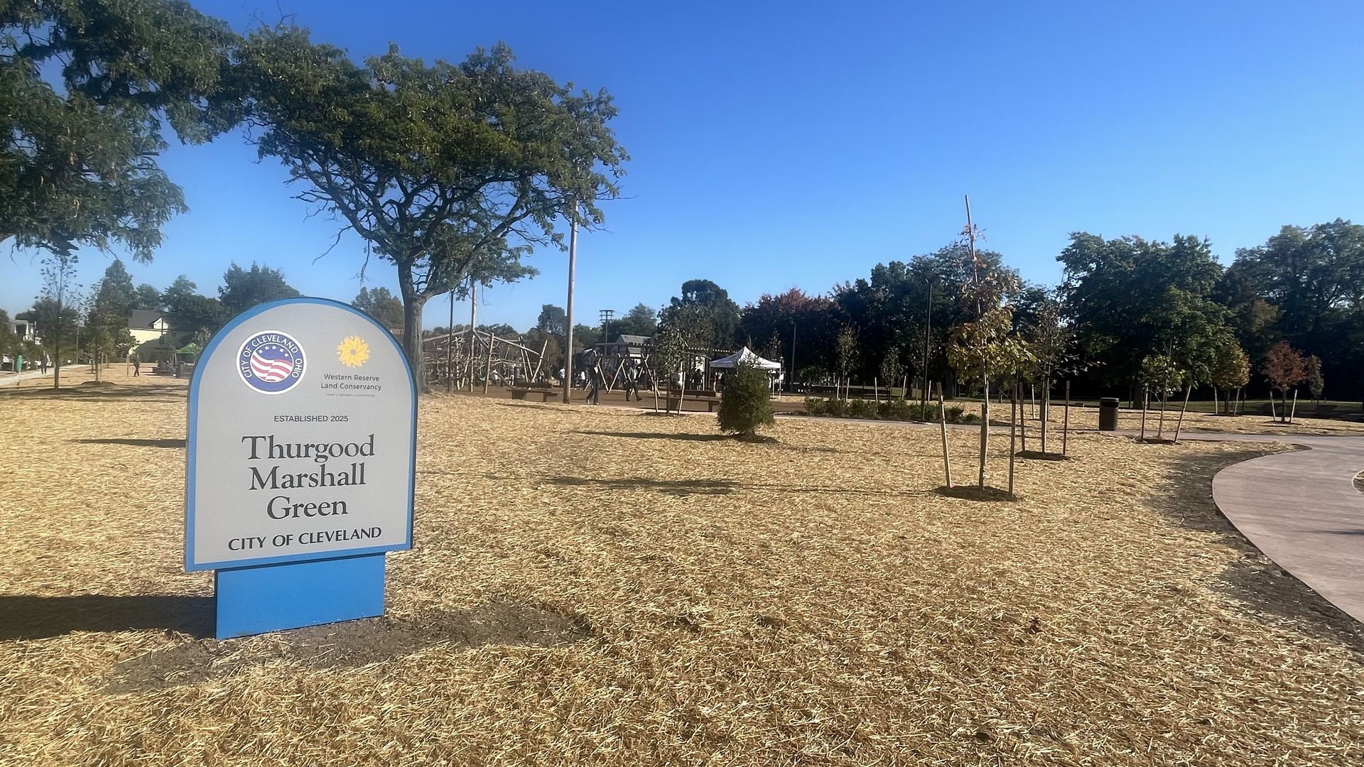 Thurgood Marshall Green park in Cleveland under clear blue sky, with a sign marking the park, trees, a curved sidewalk, and a playground area in the distance.
