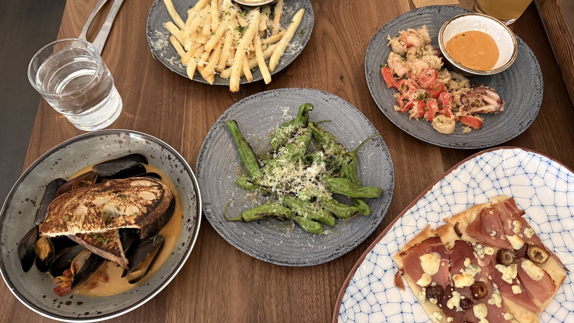 Top-down view of a wooden table with a spread: fries with cheese dip, blistered peppers, a seafood dish with orange sauce, mussels, a meat-and-cheese flatbread; plus water and a yellow drink.