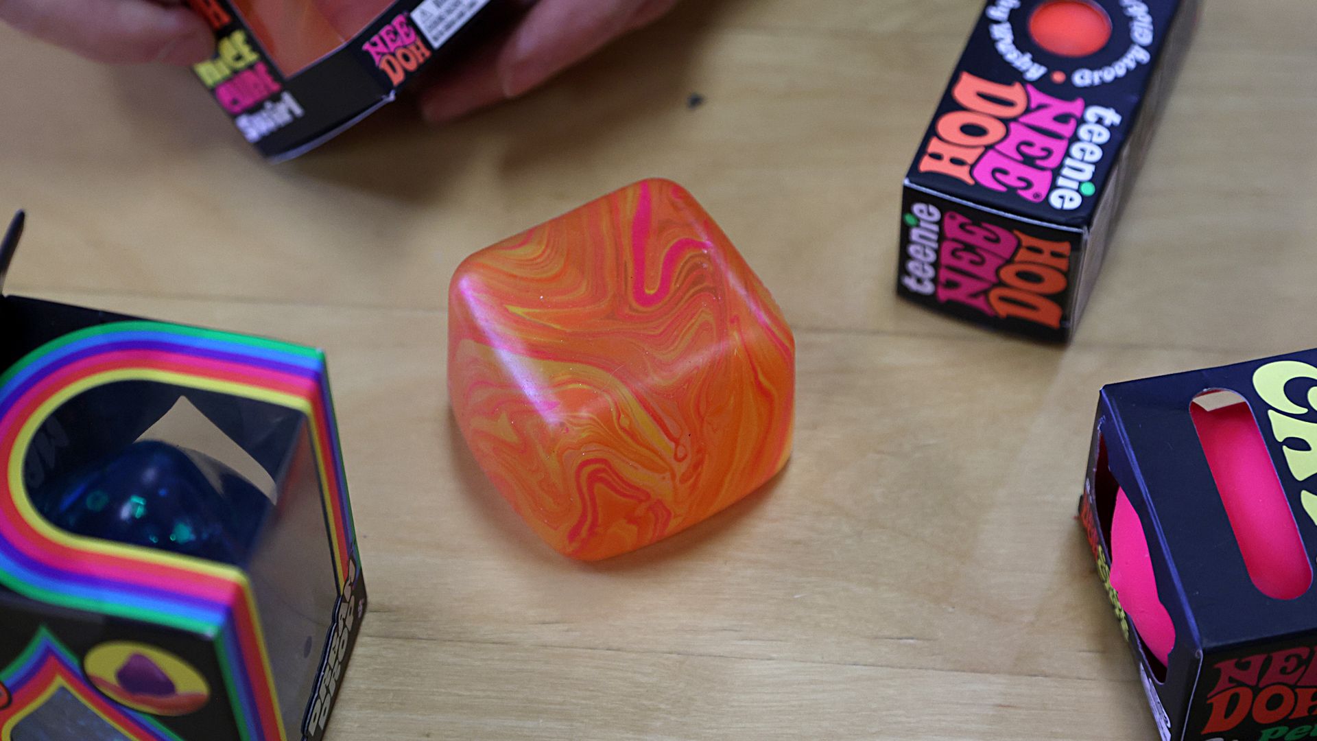 Boxes of NeeDoh sensory toys surround an unboxed orange and yellow squeezable NeeDoh cube on a table, while a person's hands hold a NeeDoh box in the background.