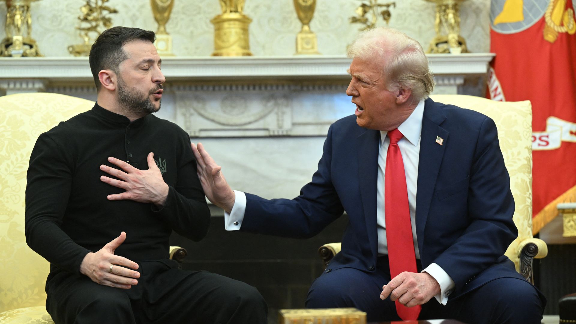 Two men seated and conversing in a formal room with golden decorations. One man, in a black outfit with a beard, gestures with his hand on his chest. The other, wearing a blue suit and red tie, reaches out with his hand.