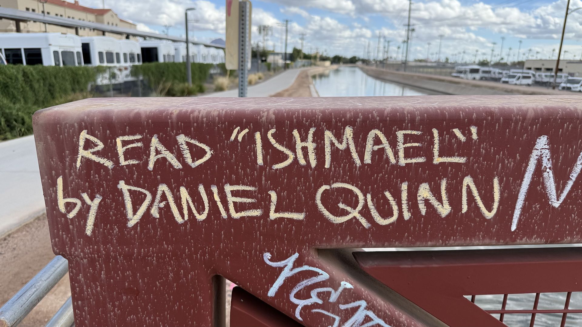 Red railing with yellow chalk graffiti that reads: Read "Ishmael" by Daniel Quinn, overlooking a canal that's running alongside buildings and white buses.