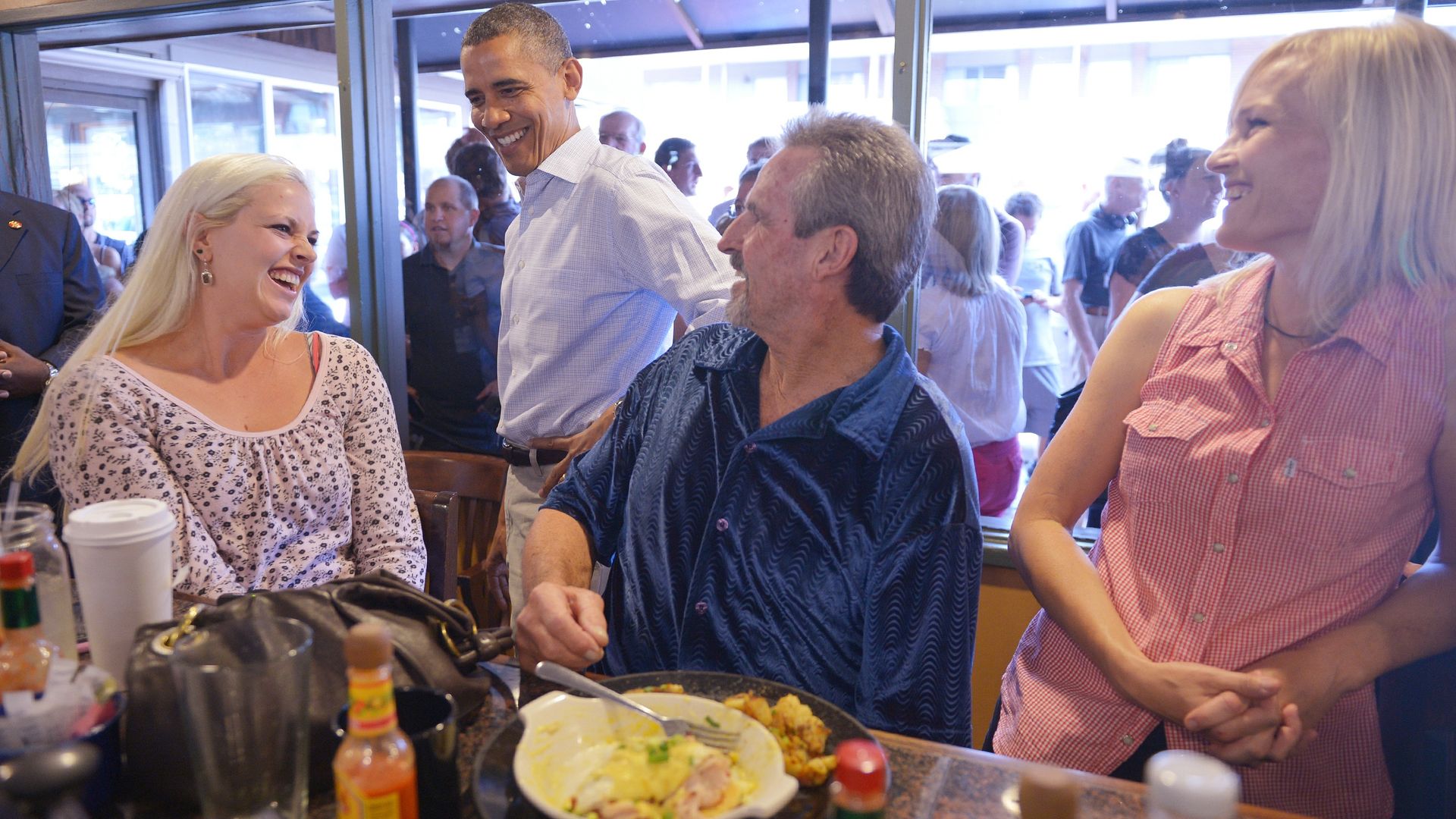 President Barack Obama chats with diners in a busy restaurant; a blonde woman in floral sits left, a gray-haired man in blue sits nearby, and a woman in pink plaid smiles on the right. Food on the table.