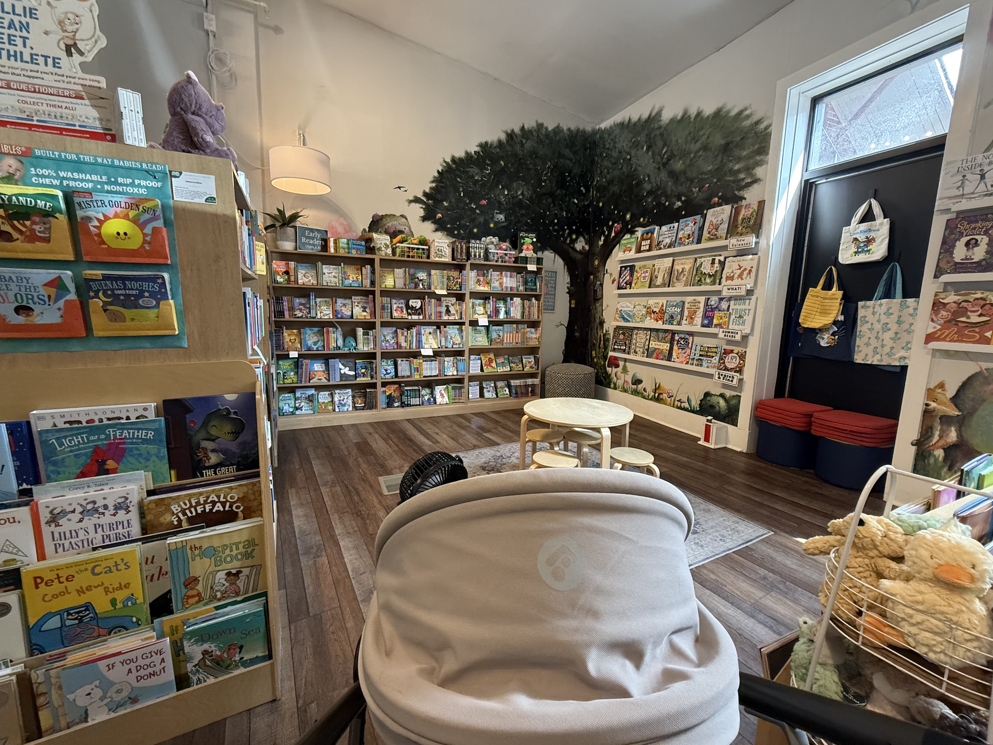 Cozy children's bookstore with colorful books on shelves, a large tree mural, plush toys, round tables, and tote bags by a doorway near a bright window.