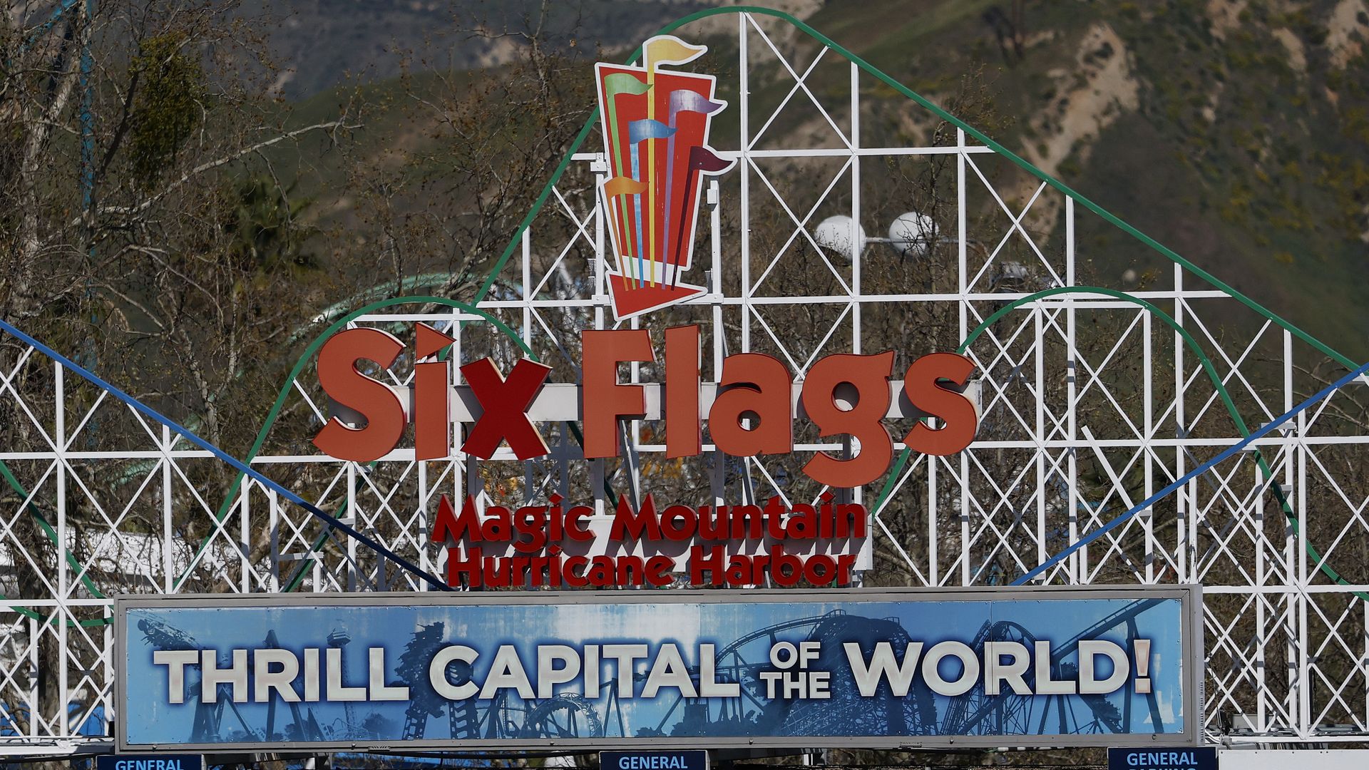 Entrance sign for Six Flags Magic Mountain and Hurricane Harbor, with a colorful flag logo atop a white lattice arch, set against hills; blue panel proclaims "Thrill Capital of the World".