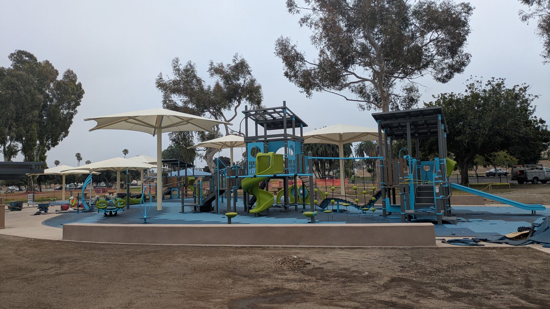 Playground with blue and green equipment on blue safety mats, white umbrellas, and tall trees under an overcast gray sky.