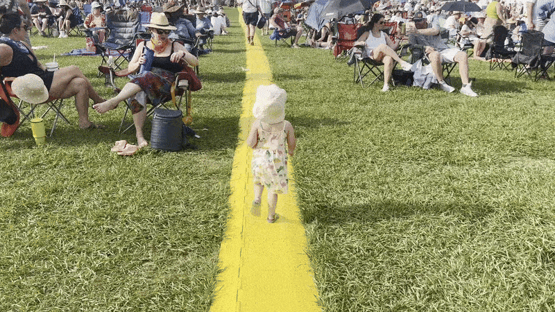 A small child runs along a yellow strip of plastic in the middle of a large festival audience.