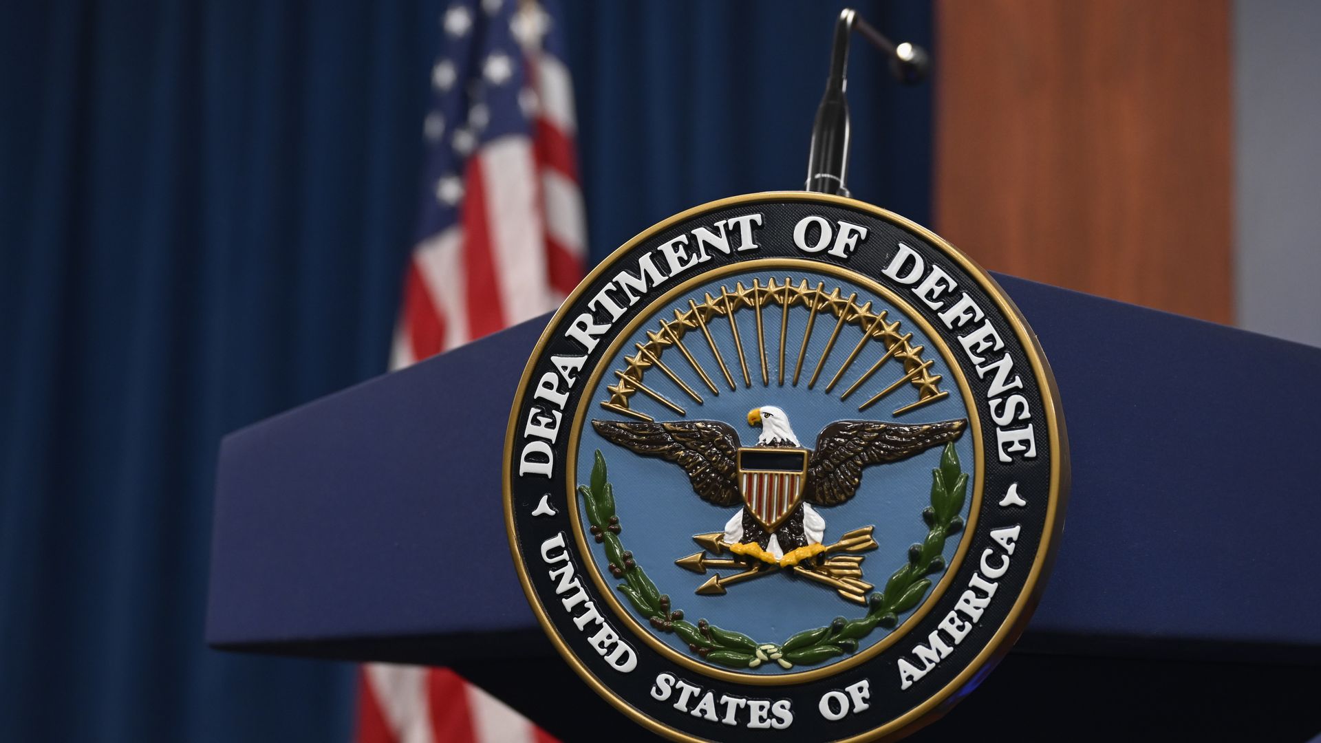 The Department of Defense logo, an eagle, is seen on a podium with a microphone. An American flag is behind it.