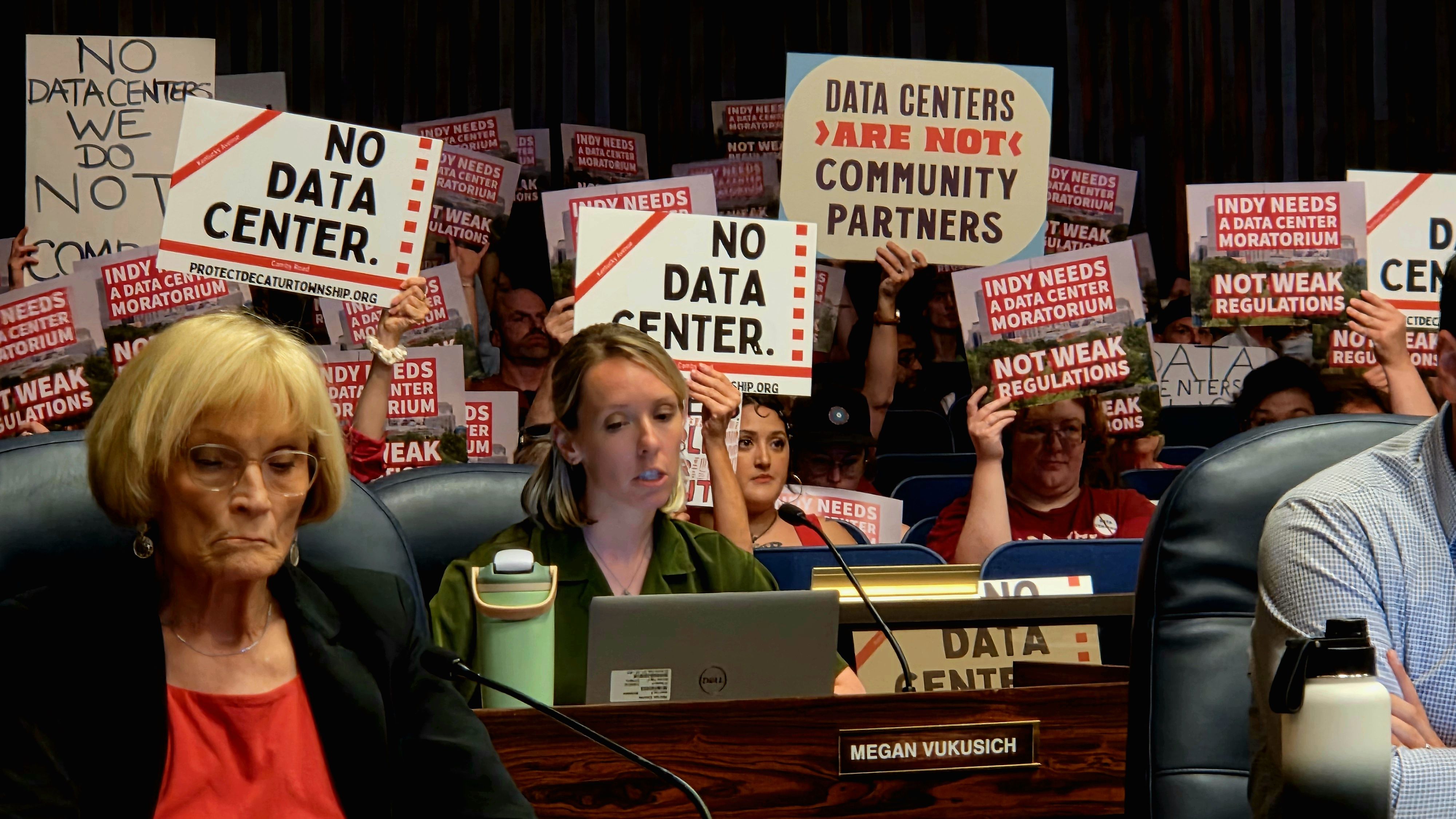 Crowded meeting with protesters holding signs reading "NO DATA CENTER" and "DATA CENTERS ARE NOT COMMUNITY PARTNERS." Foreground shows a woman in green at a desk with a mic and laptop.