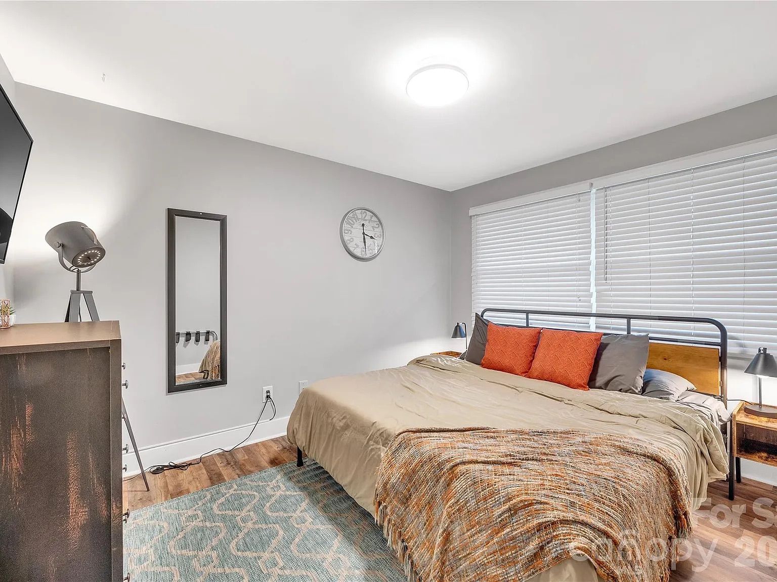Cozy modern bedroom with a metal bed frame, beige bedding and orange pillows, blue patterned rug, gray walls, white blinds, dresser with a metal lamp, tall mirror, and round clock.
