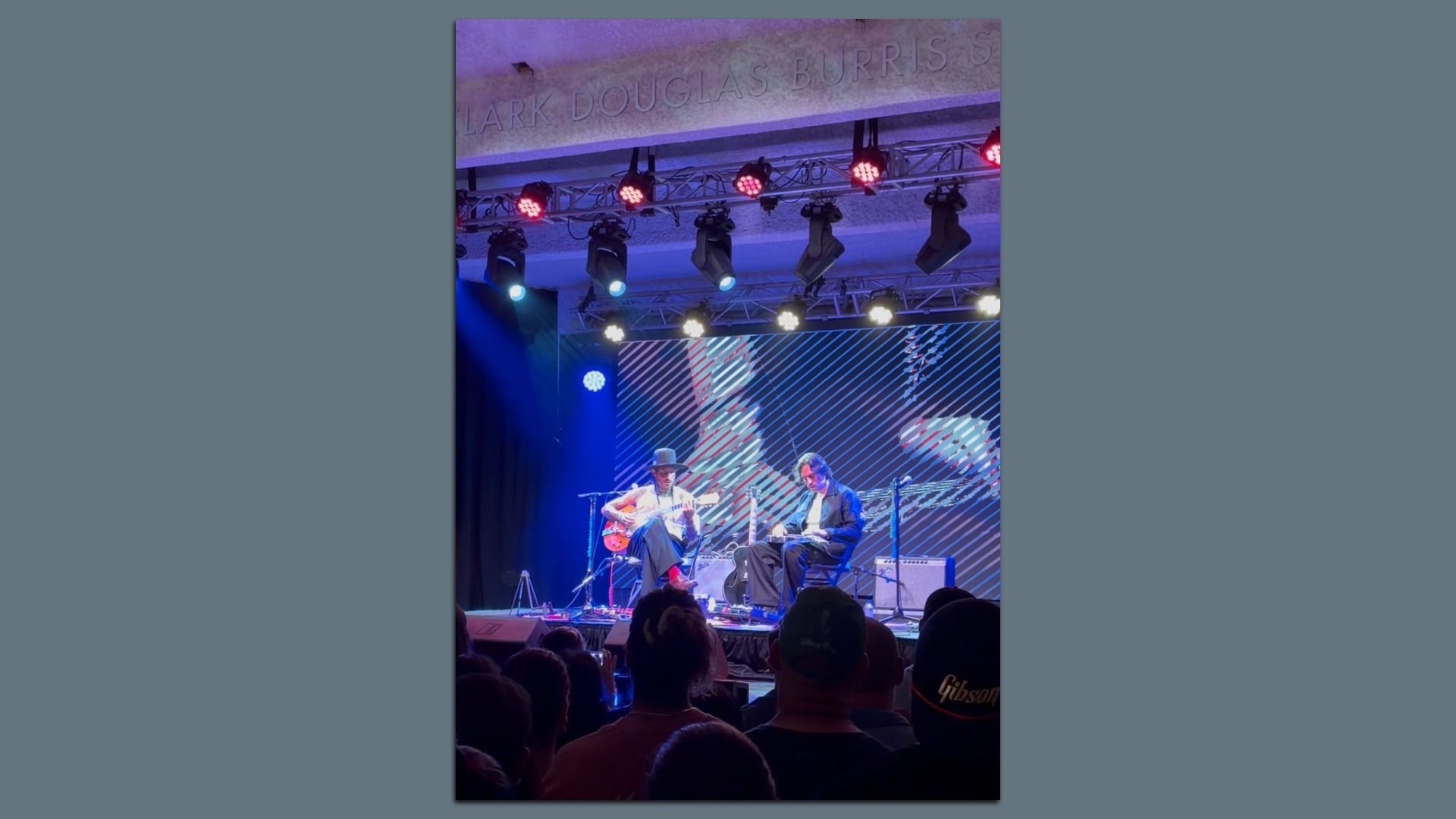 Two musicians seated on stage playing guitars under purple and blue lights, with an audience in front and a screen with abstract visuals behind them.