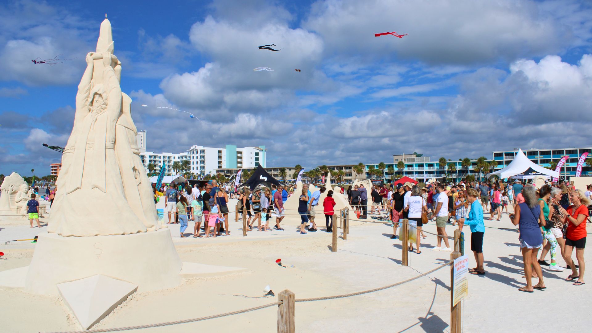 Crowded beach event with large detailed sand sculptures, people walking and observing, colorful kites flying, blue sky with clouds, and buildings in the background.