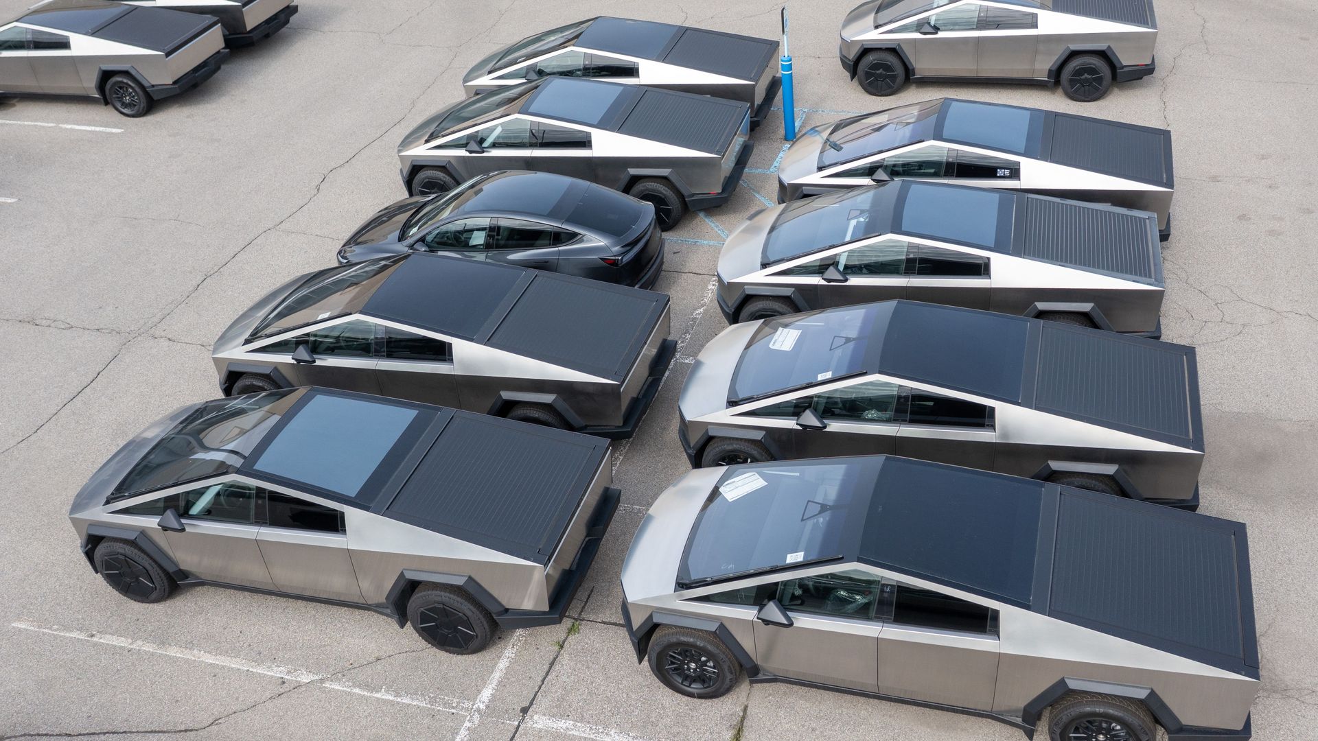 An aerial view of a parking lot featuring multiple futuristic, angular vehicles with stainless steel exteriors. These vehicles have a distinct geometric design and are parked in an organized formation. One traditional car is parked among them, standing out due to its different shape and color.
