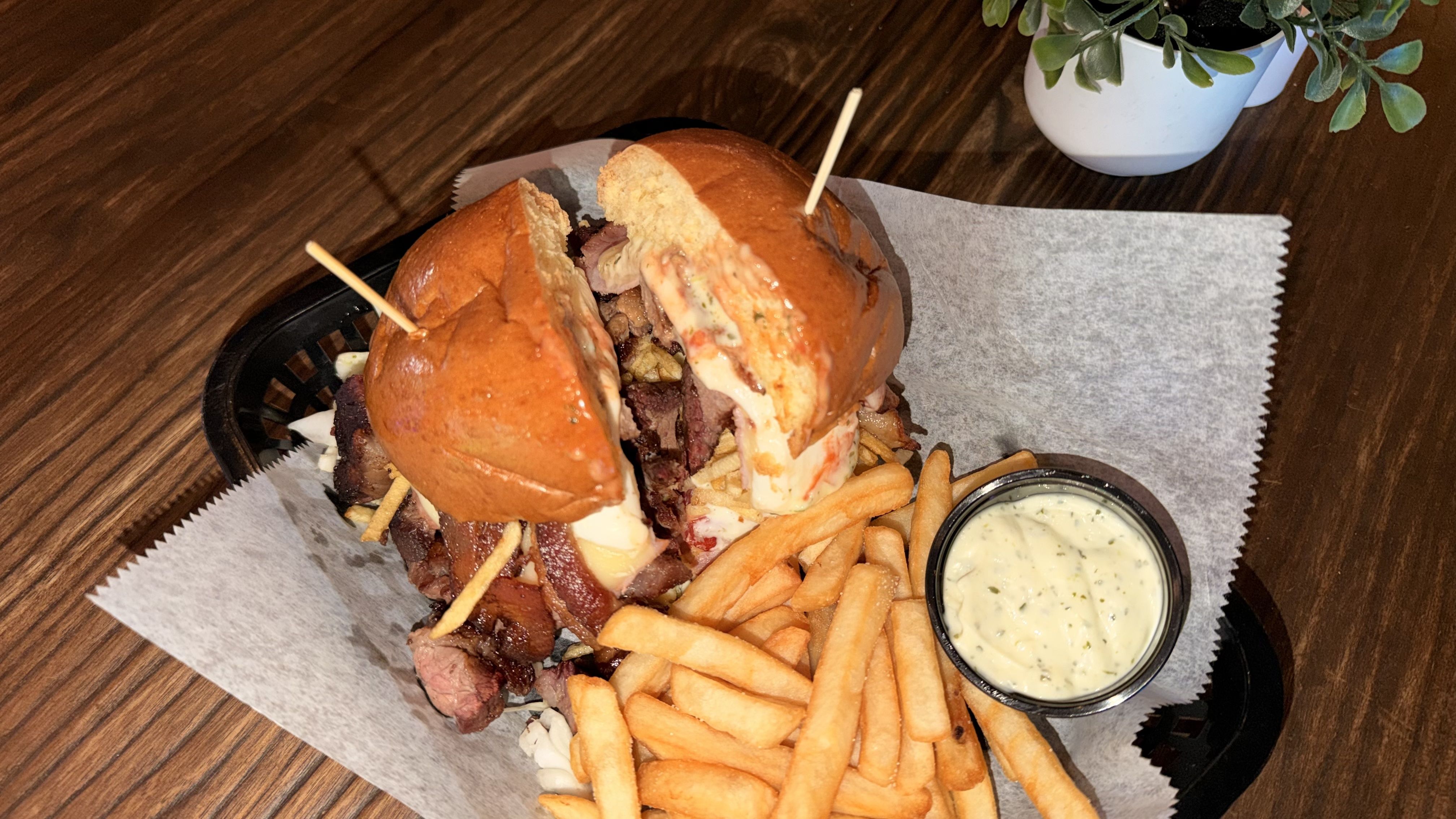 Overhead shot of a loaded burger with melted cheese and pulled beef, split buns held by toothpicks, with fries and a creamy dip on parchment on a dark wooden table; a potted plant in the background.