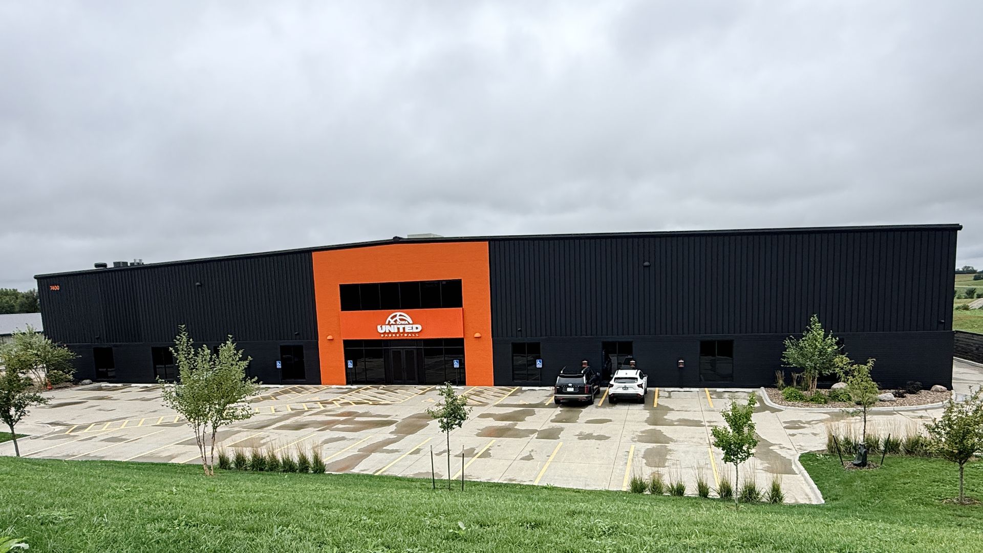 A long black industrial building with a bright orange entry panel and a white United Basketball logo; a parking lot with several cars in front, trees, and a cloudy gray sky.