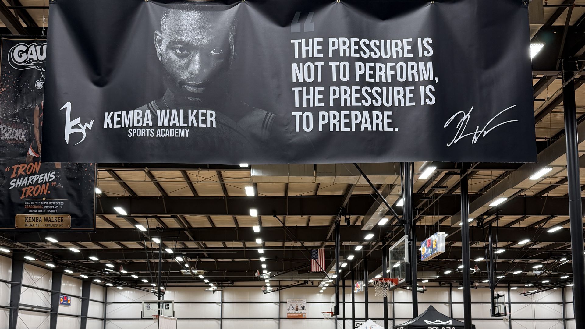 Indoor basketball gym with a large black banner displaying a grayscale portrait of a male athlete and the quote "The pressure is not to perform, the pressure is to prepare." The banner also reads "Sports Academy."