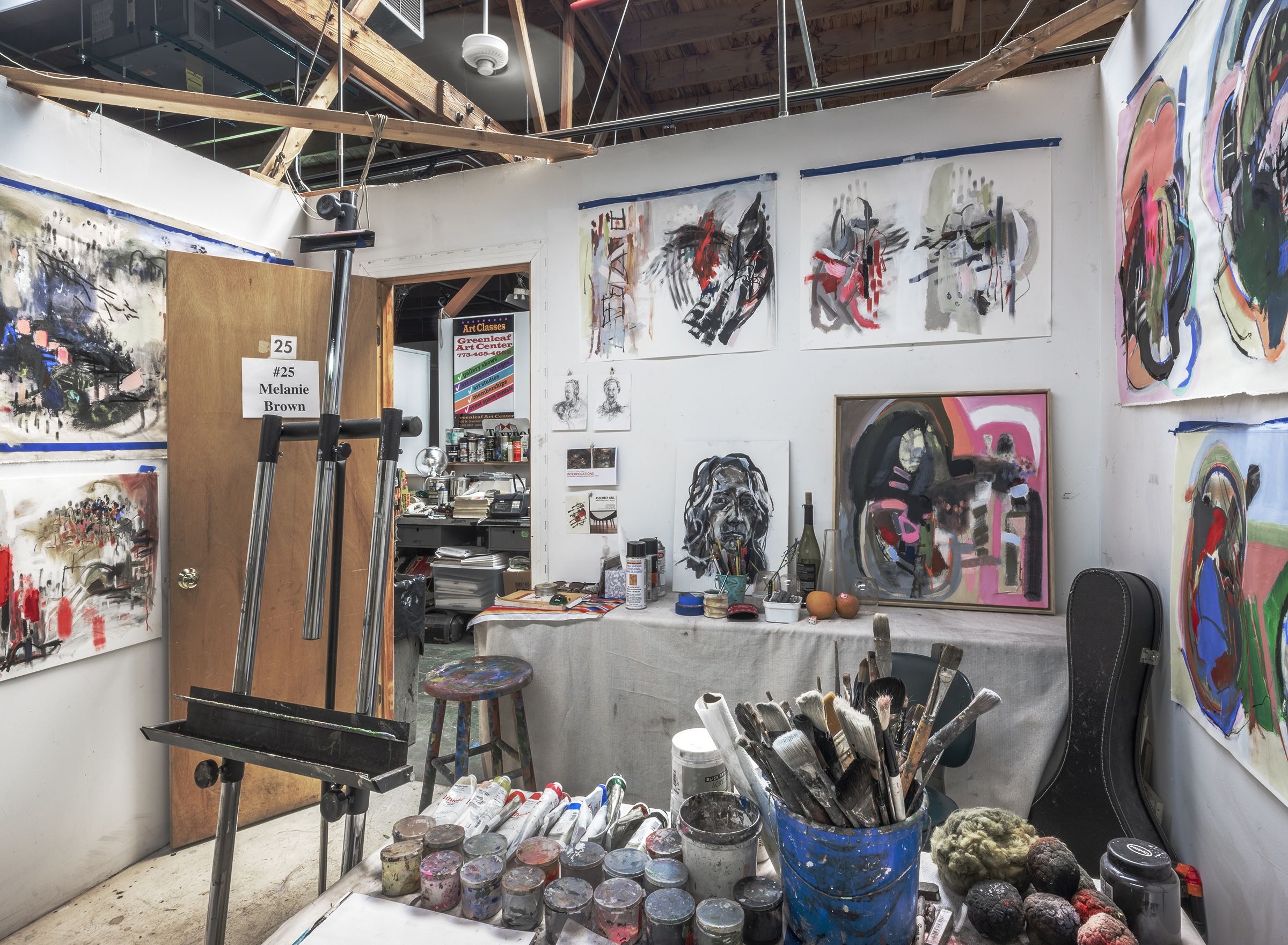A small studio space with an empty easel and paintings and drawings on the wall and a container with paint brushes.