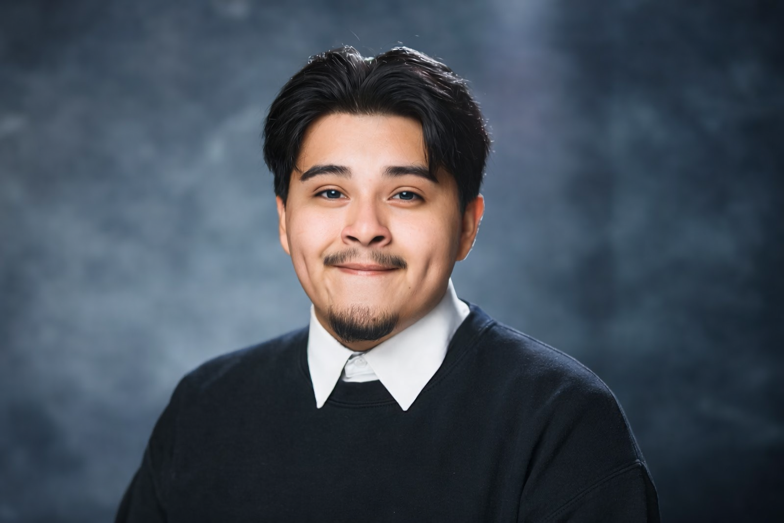 Head-and-shoulders portrait of a young man with black hair and a light mustache/goatee, wearing a white collared shirt under a dark sweater, smiling softly against a blue smoky studio backdrop.