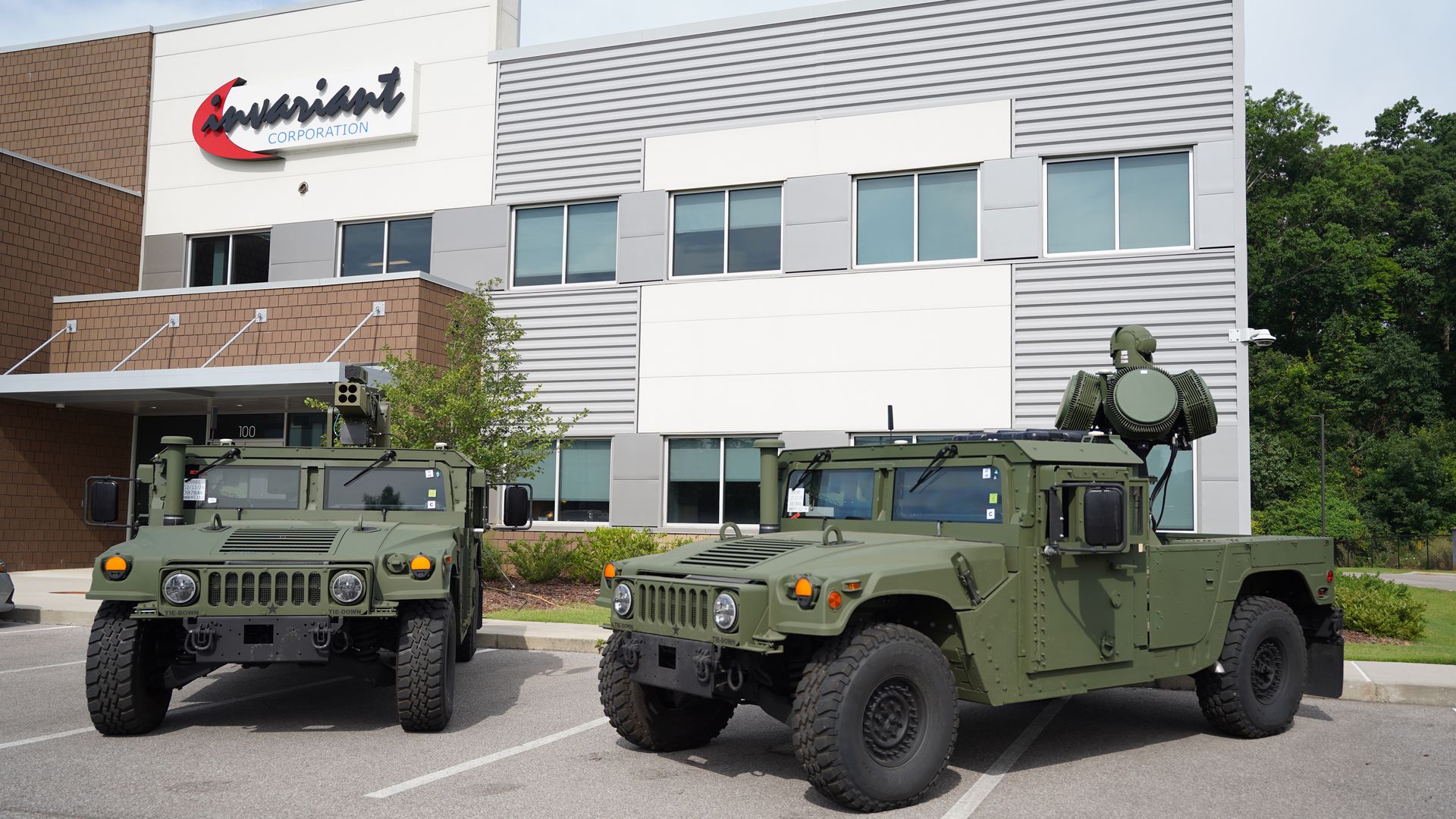 Exterior of a modern office building with a red and black "Invariant Corporation" sign; two large green military trucks with turrets are parked in front, with trees in the background.