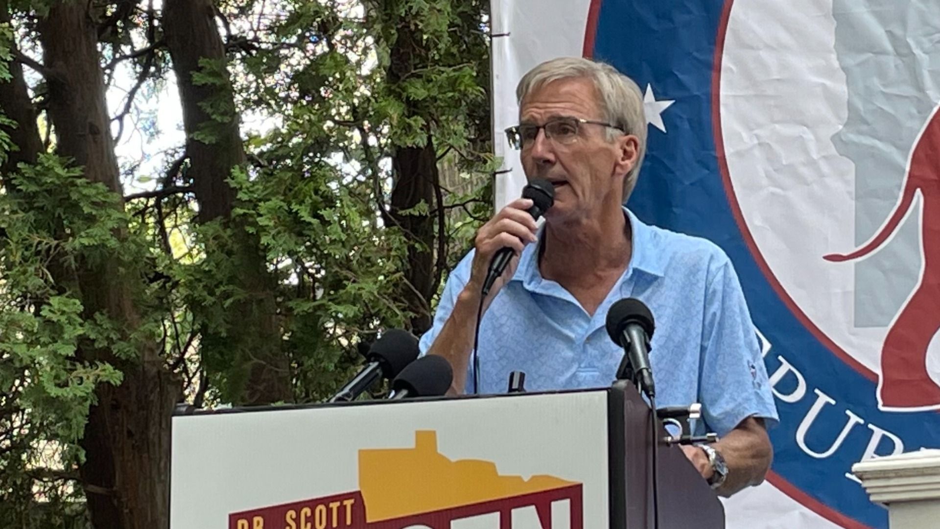 Scott jensen speaking into a microphone at podium with D.R. Scott sign, green trees behind him, and a partially visible Republican Party banner in background.