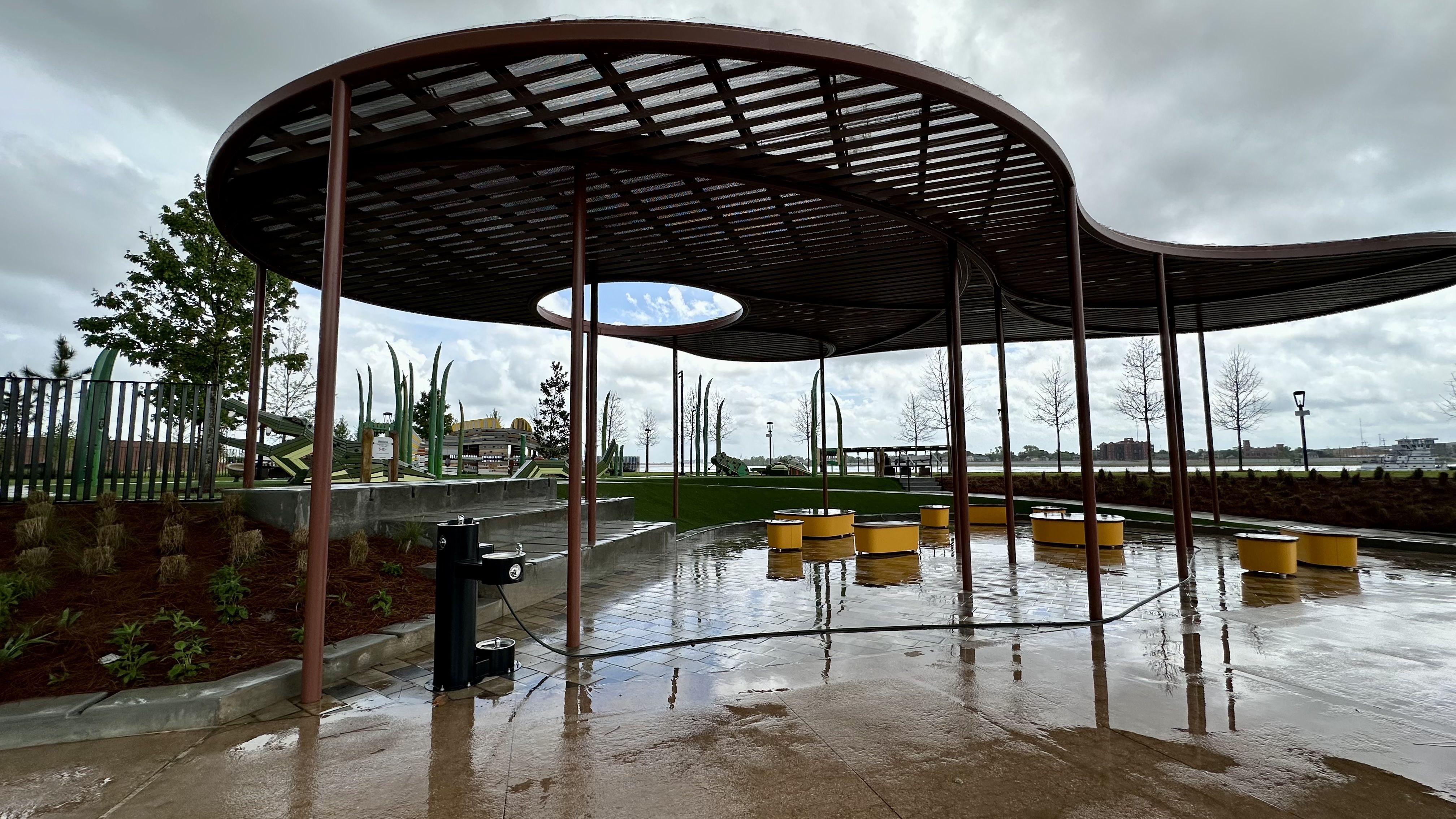 Open-air curved brown pavilion with tall posts over a wet concrete plaza, yellow circular stools, a playground in the background, and a cloudy waterfront view with distant buildings.