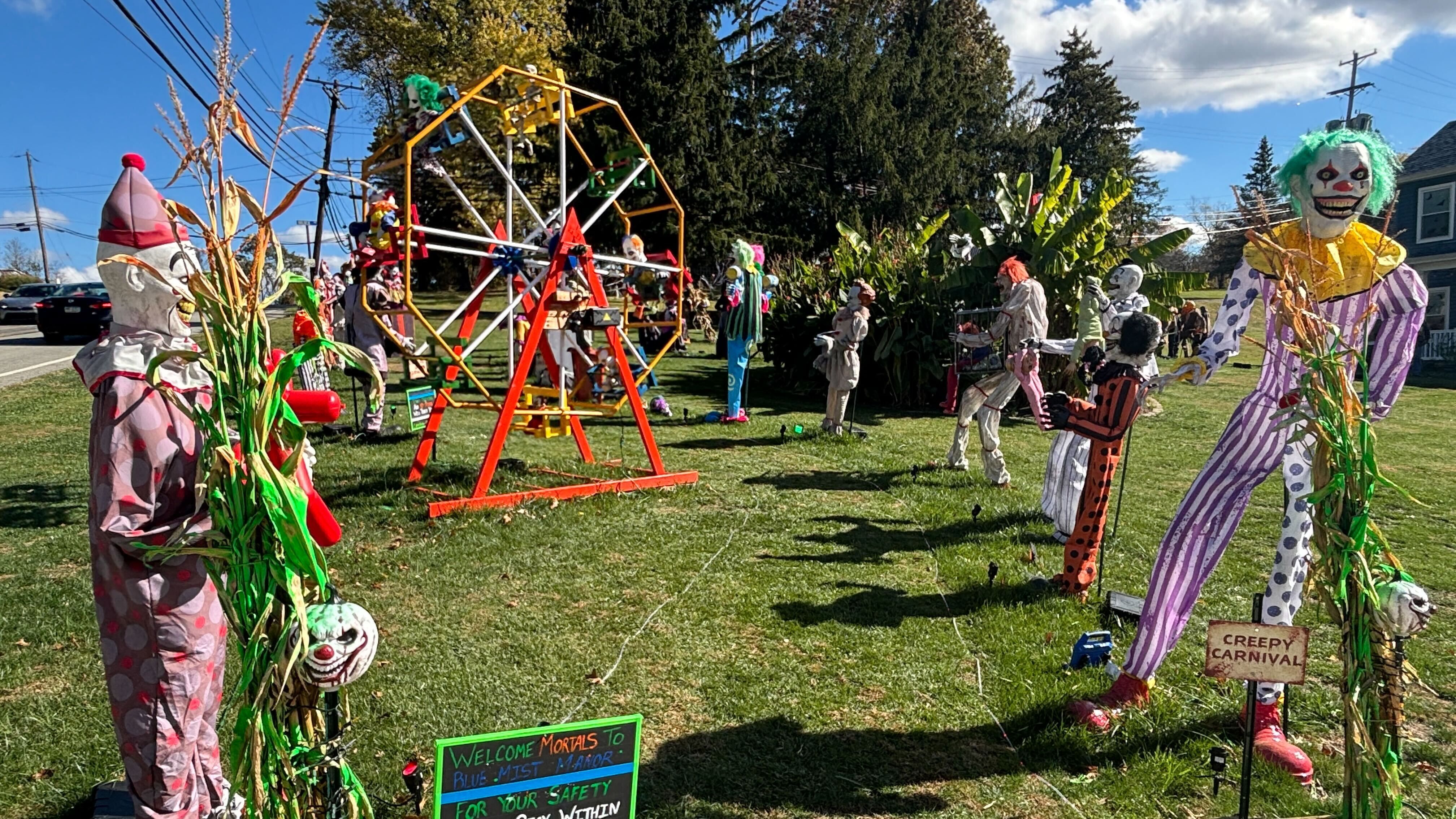 Outdoor Halloween display with large creepy clown figures, a small Ferris wheel with clown dolls, and scary signs reading "Creepy Carnival" on green grass under blue sky.