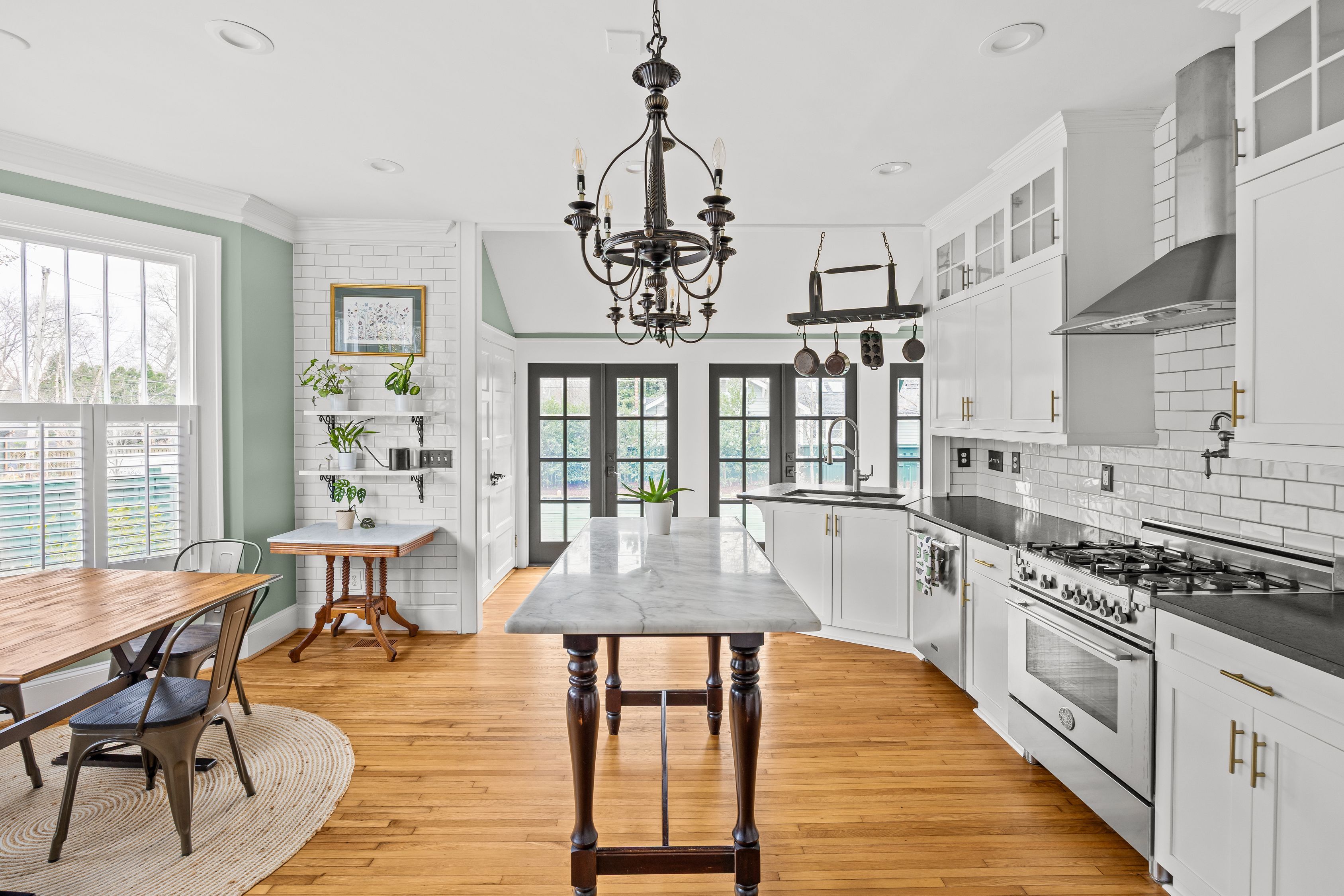 Bright kitchen with white cabinets, black countertops, subway tile backsplash, wooden floor, central marble island, black chandelier, hanging pots, and large windows and doors letting in natural light.