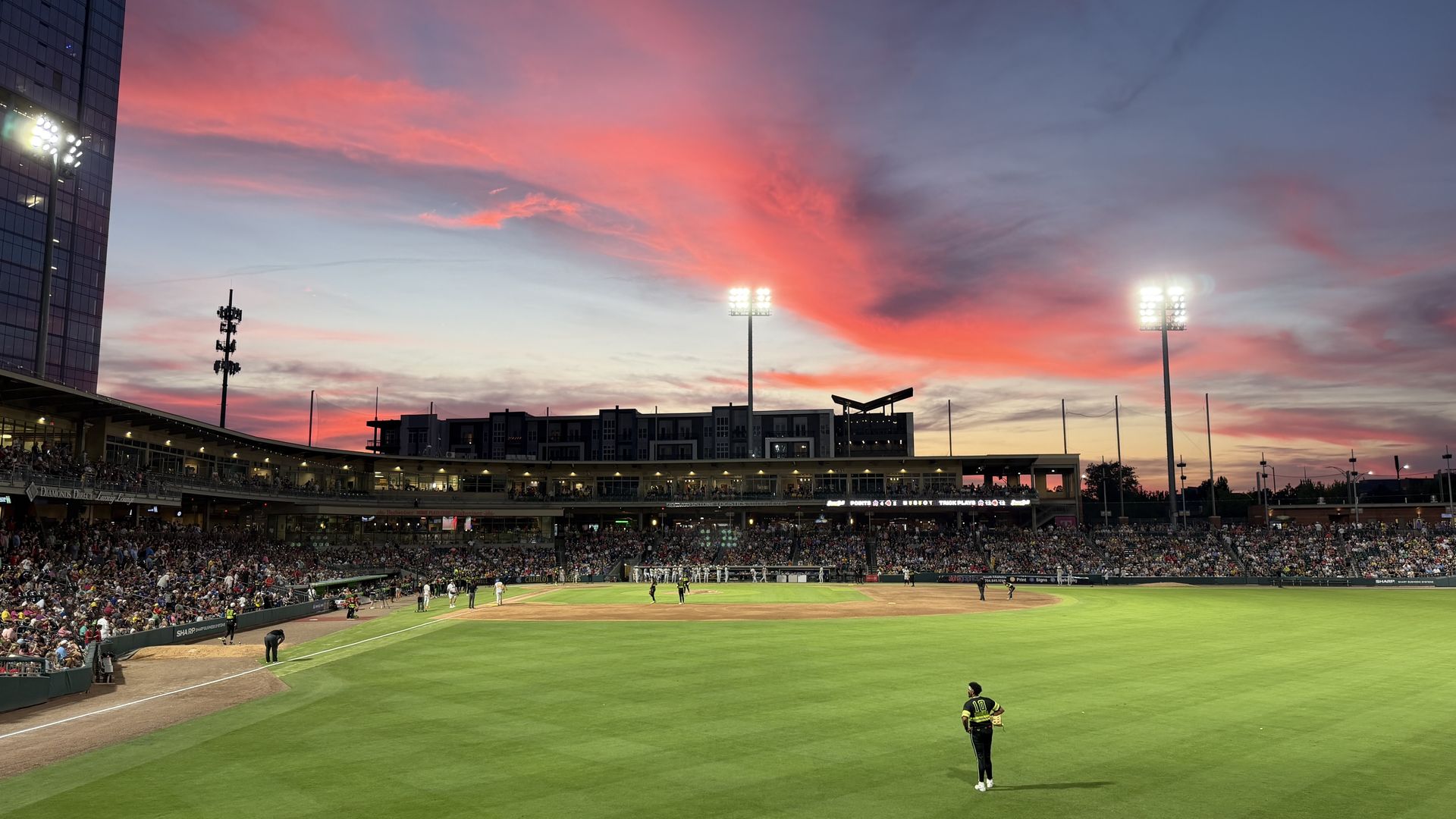 Baseball stadium at sunset with pink and orange clouds, bright floodlights, a full crowd in the stands, players on the infield, and a tall glass building on the left against a purple evening sky.