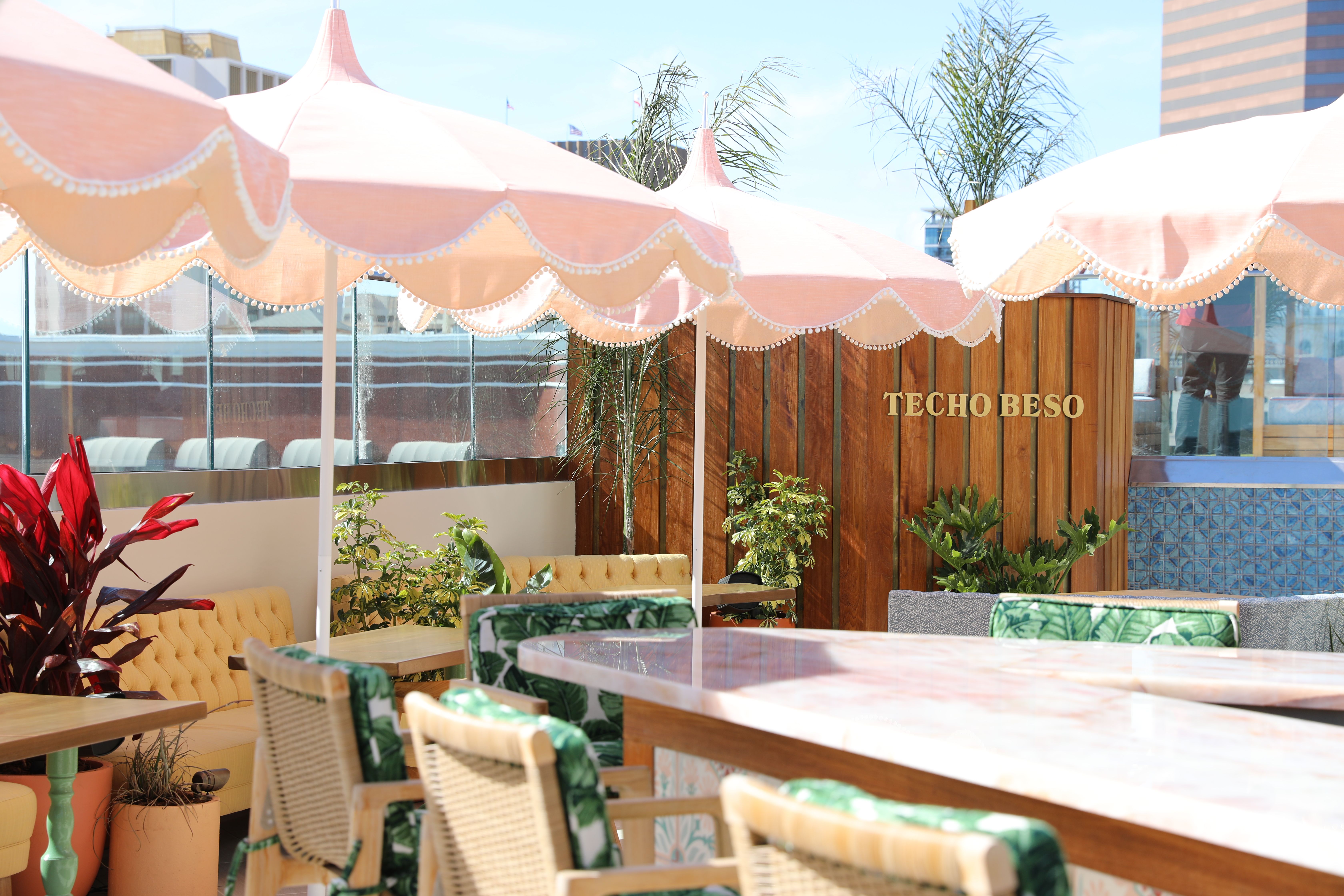 A rooftop bar with coastal decor and furniture including pink umbrellas and palm-printed chairs.