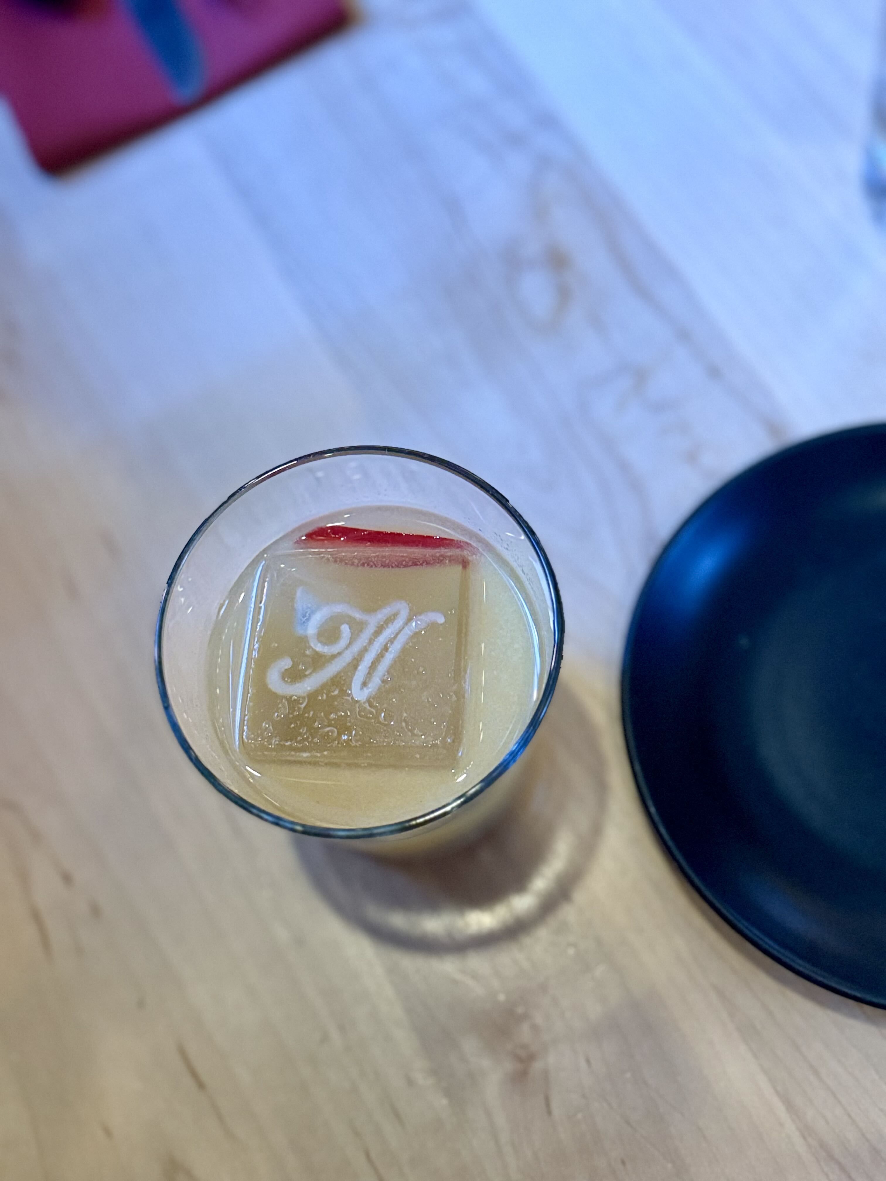 Pale yellow drink in a glass with a large ice cube etched with a white M, on a light wooden table; a dark blue object is nearby.