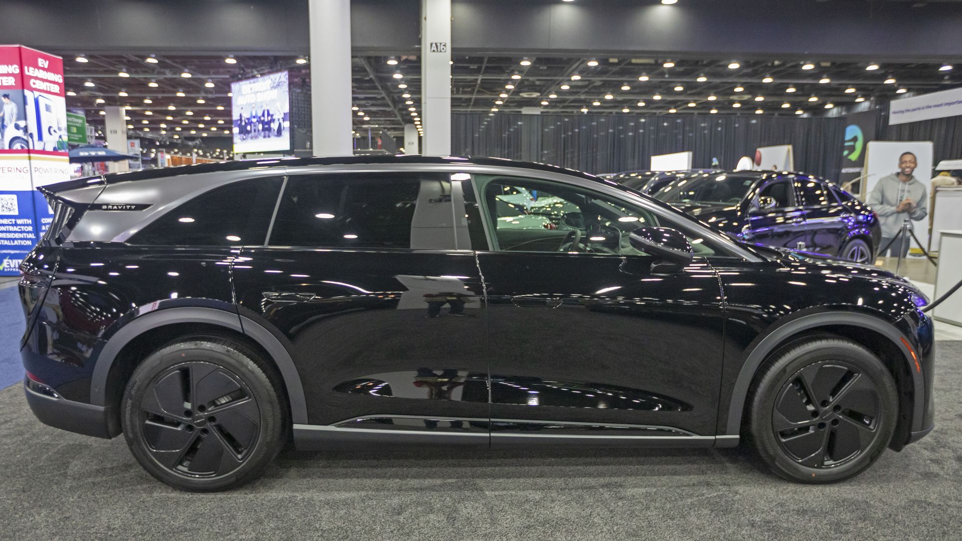 Glossy black SUV dominates a busy indoor car show, with large alloy wheels and a "GRAVITY" badge. Bright overhead lights, other cars, and exhibition booths in the background.