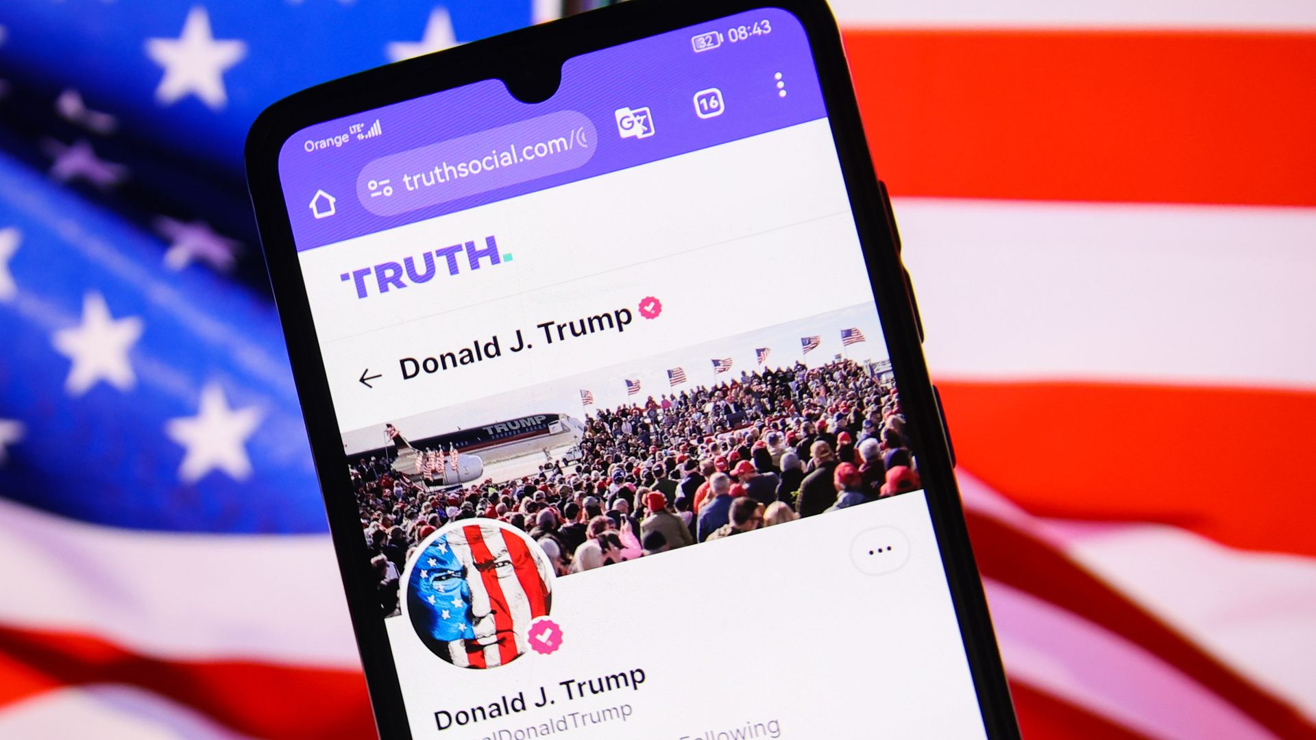 A smartphone displaying Donald Trump’s Truth Social profile is pictured in front of a blurred American flag.