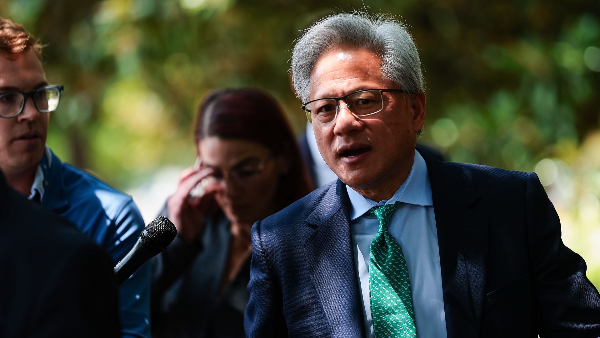 A person with gray hair and glasses in a blue suit and green tie speaks into a microphone outdoors, with blurred people and green foliage in the background.