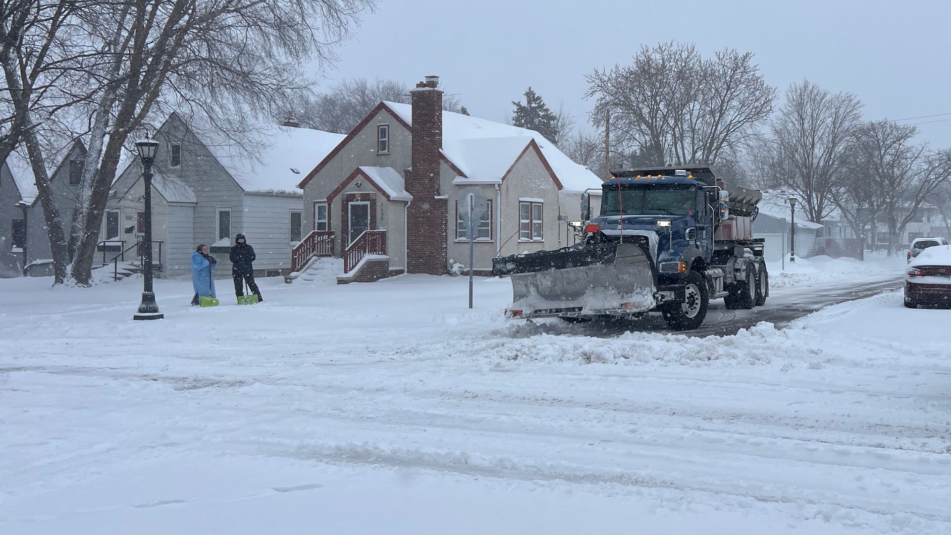 Blue snowplow clears a snowy residential street; a second plow sits to the right. Snow-covered houses, bare trees, and parked cars blanketed in white, with two people in bright coats near a lamppost.