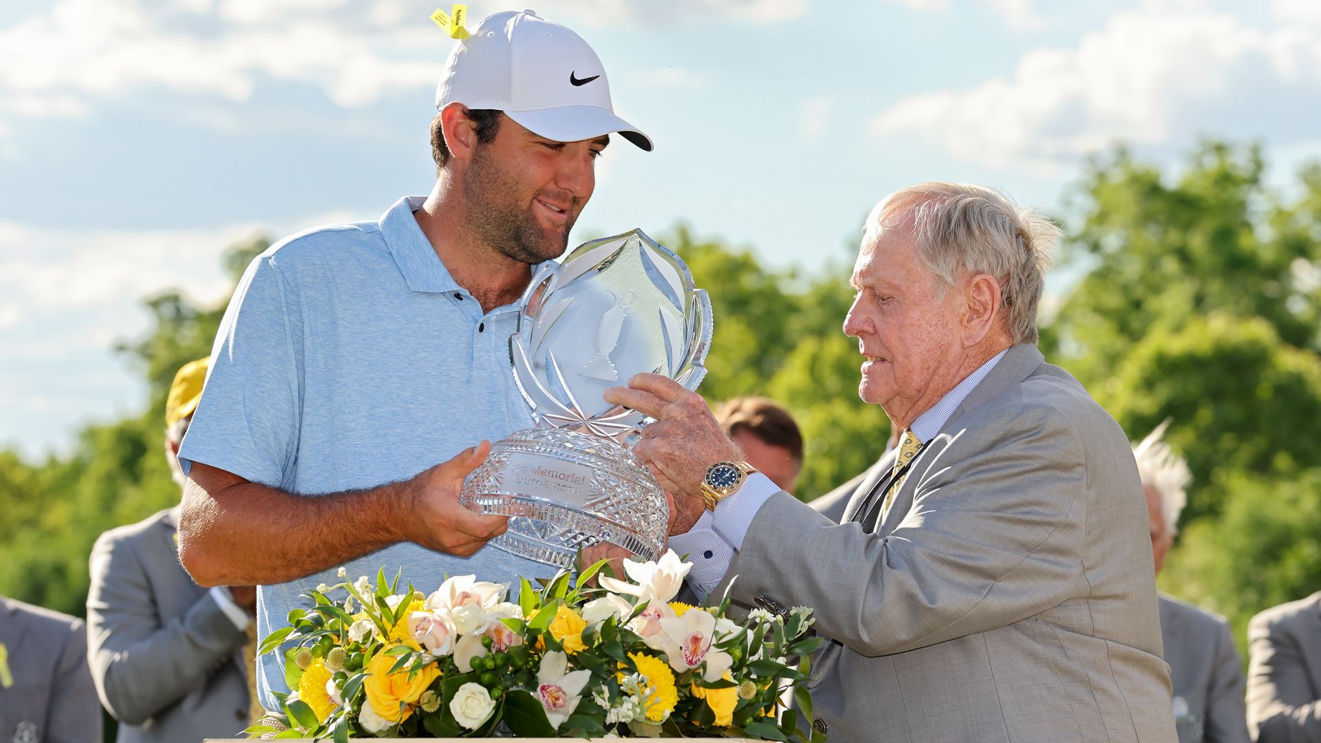 Jack Nicklaus hands a Memorial golf tournament championship trophy to winner Scottie Scheffler in front of a bed of flowers.