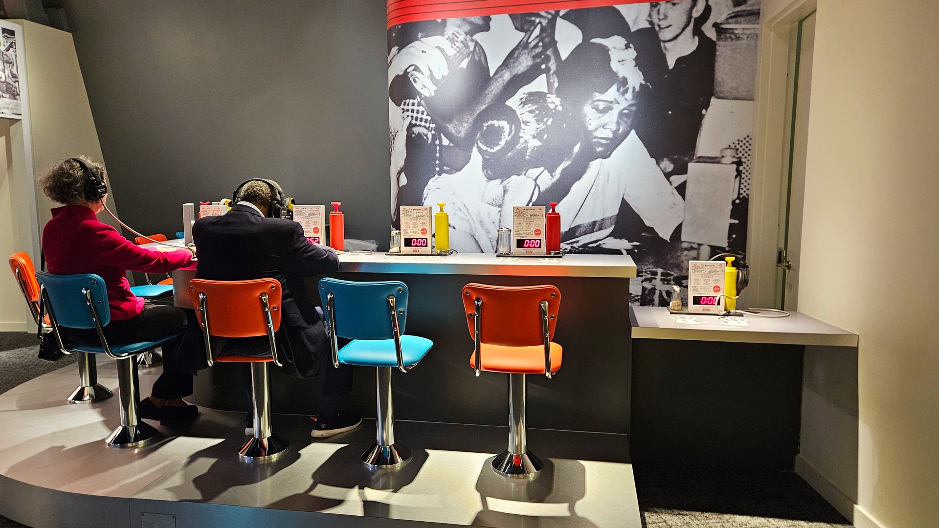 Two people sitting on colorful retro-style bar stools wearing headphones at a counter with menus and ketchup/mustard bottles, in front of a black-and-white photo wall.