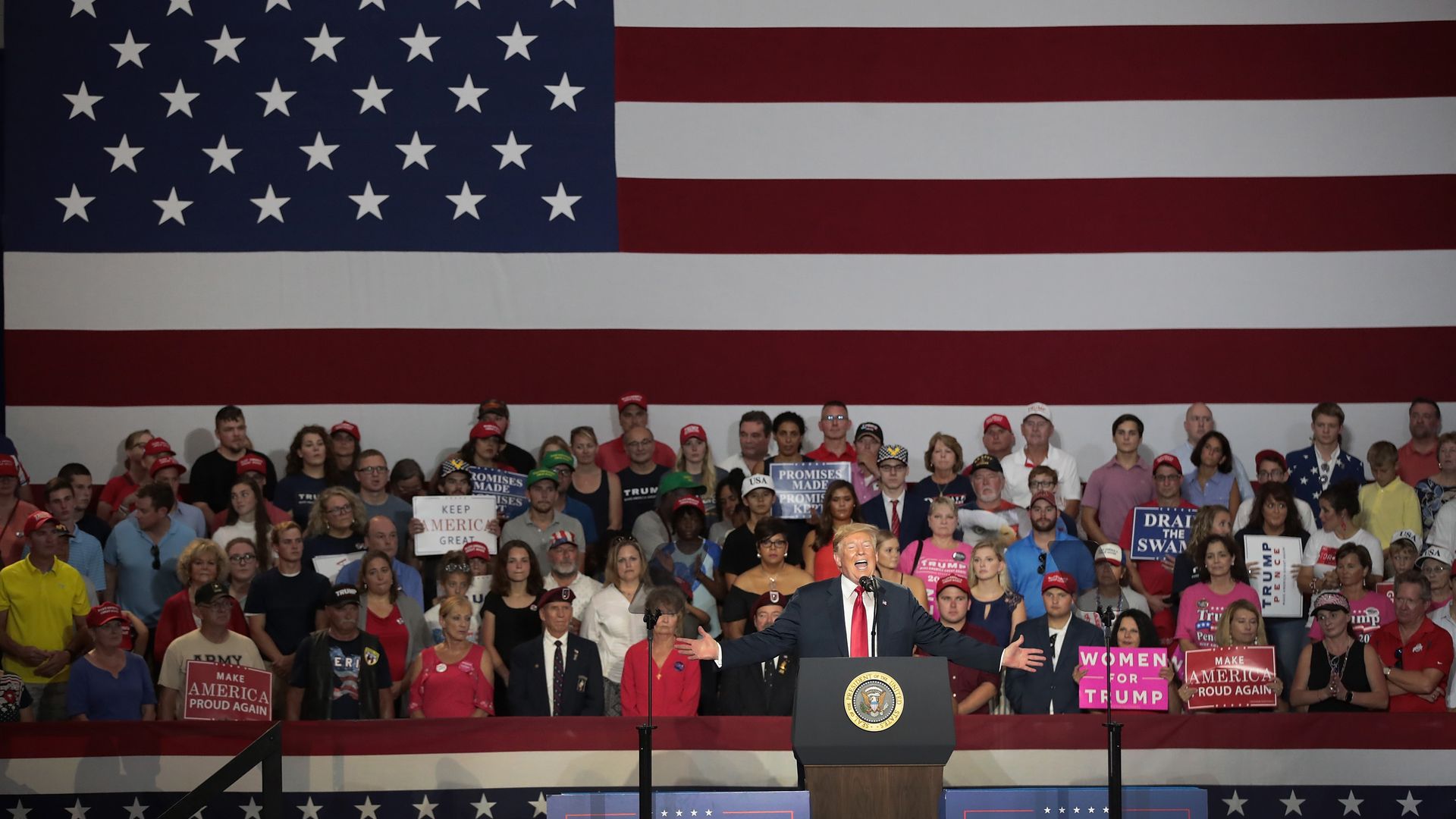 President Trump speaking in front of a crowd and an American flag backdrop at a rally.