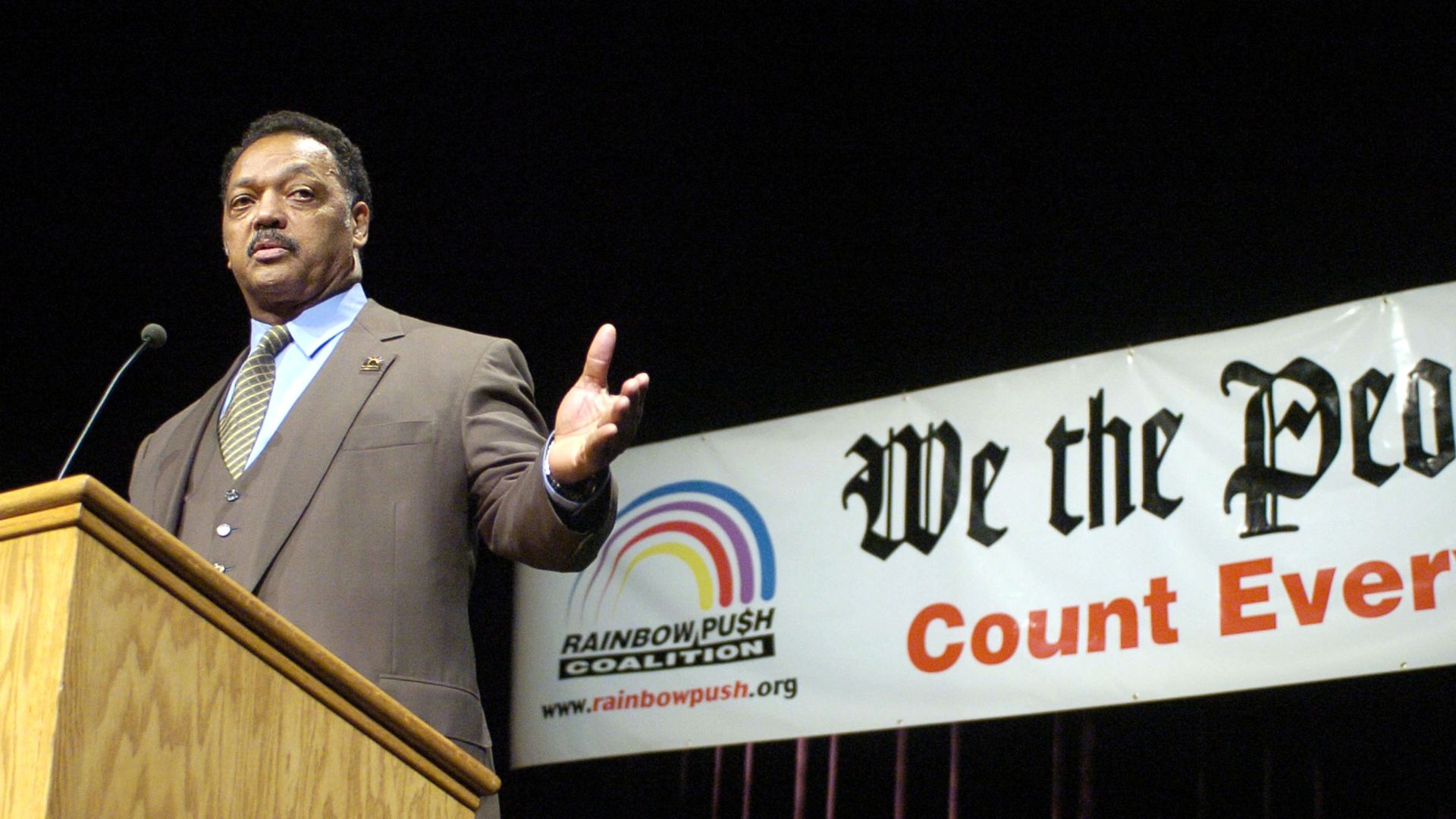The Rev. Jesse Jackson speaks at a wooden podium with a banner behind him reading "We the People Count Every Vote" and showing the Rainbow PUSH Coalition logo and website.