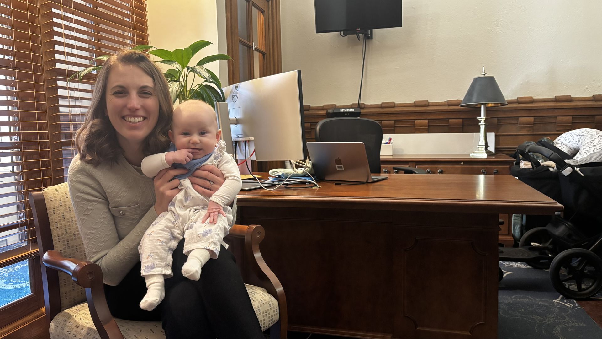 Smiling woman sits in a wooden chair, holding a baby in light clothes. They're in a warm office with a large desk, a Dell monitor, a silver laptop, a lamp, wood-panel walls, and blinds.
